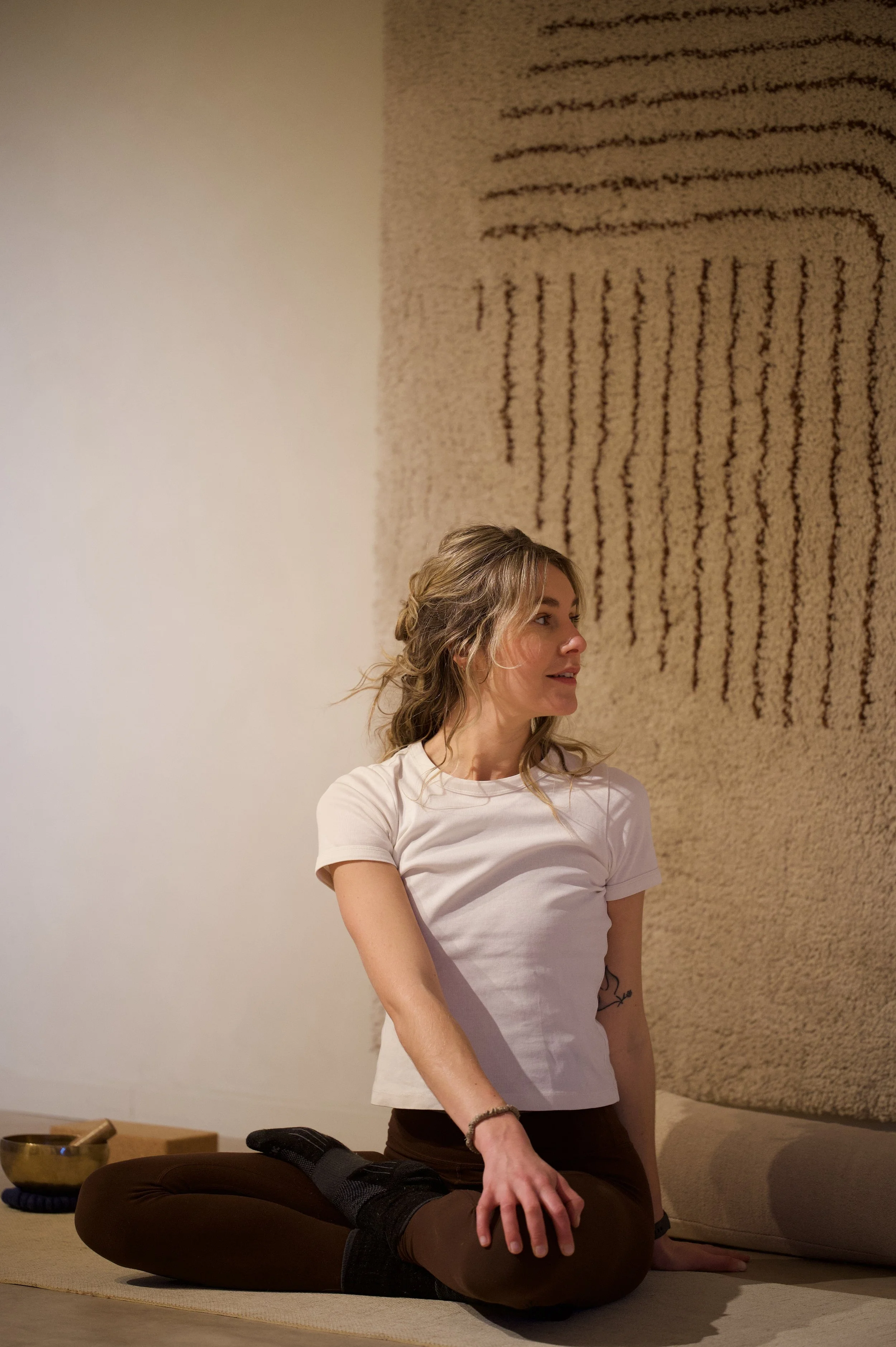 Een vrouw met blond haar in een witte T-shirt zit gekruist op de grond, met haar linkerhand op haar knie in een kalme ruimte, met een beige muur en een kunstwerk met verticale lijnen op de achtergrond.