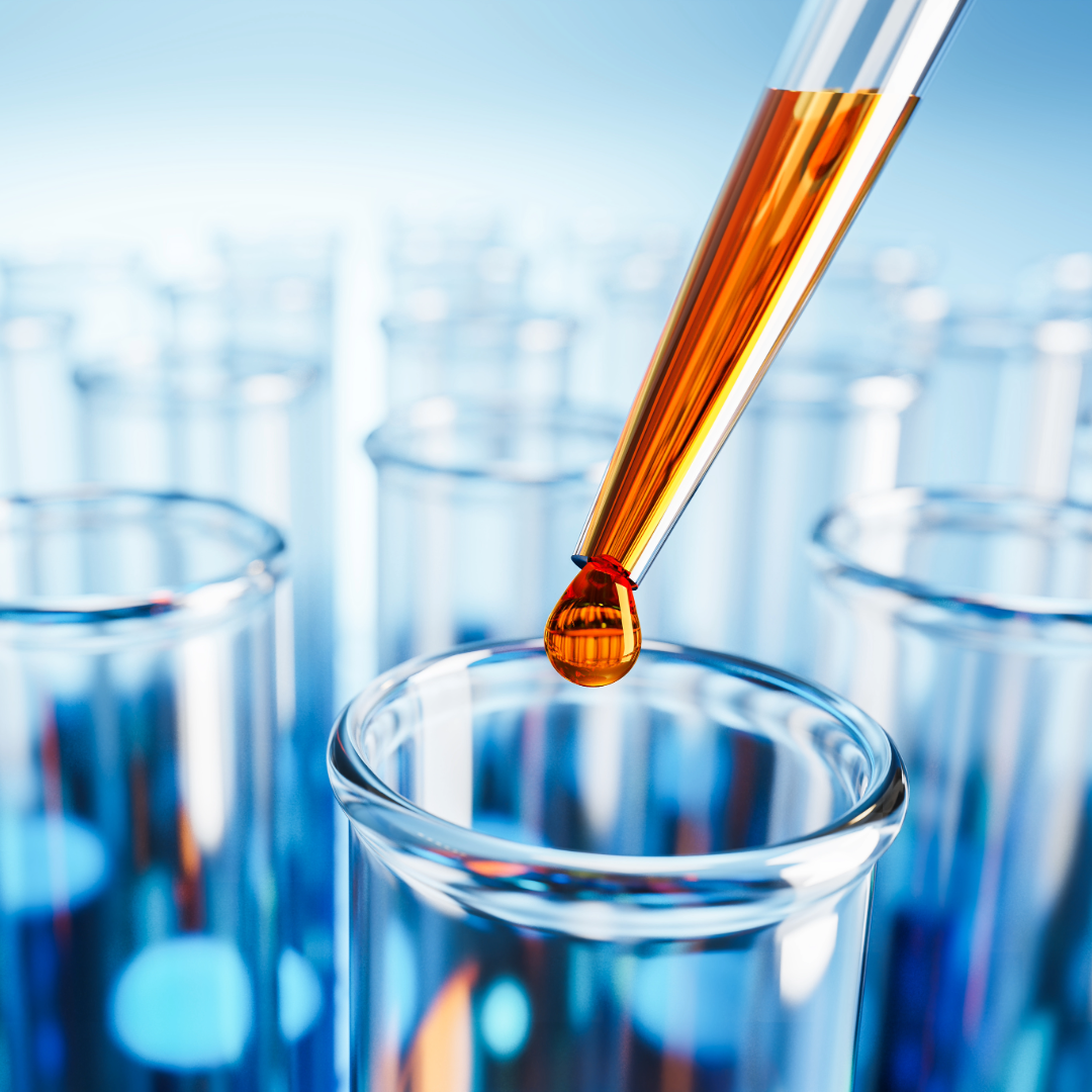 Close-up of a pipette dispensing a drop of orange liquid into a test tube in a laboratory, with several other test tubes in the background.