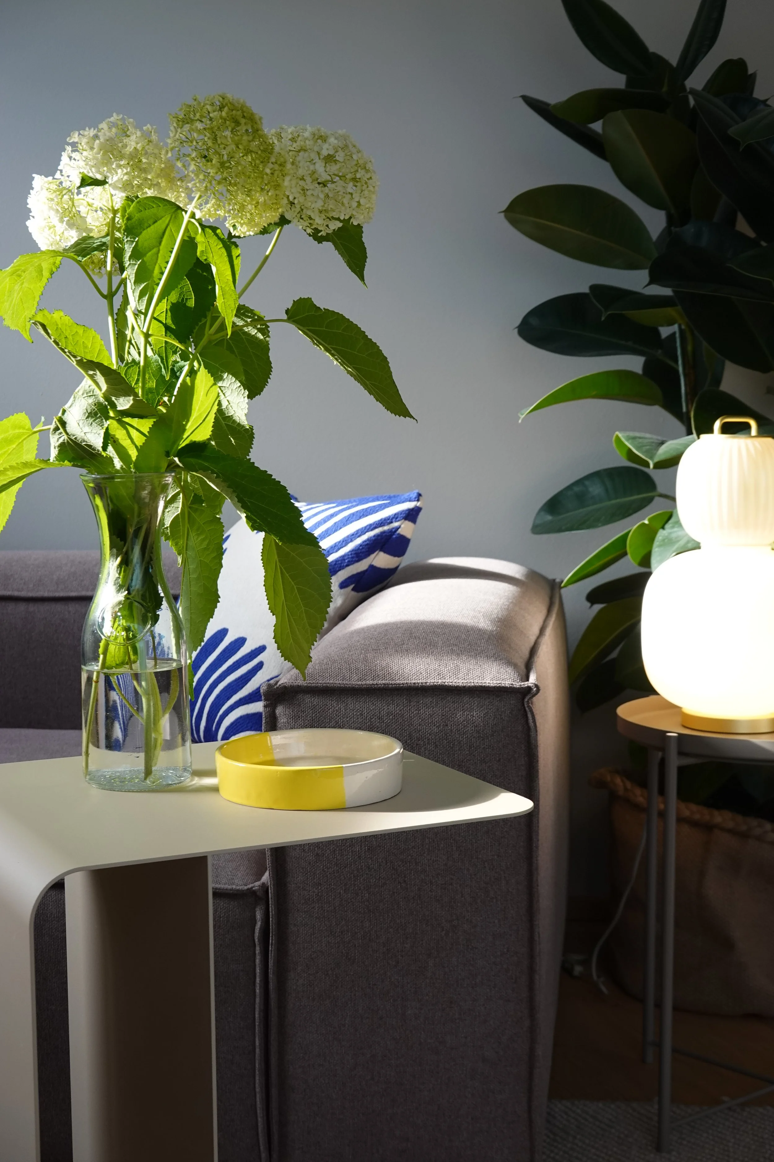 Wohnzimmer mit taupefarbenem Sofa, hellgelbem Beistelltisch, Keramikschale, Hortensien, blau-weiß gestreiftes Kissen, moderne Tischleuchte mit organischen Formen