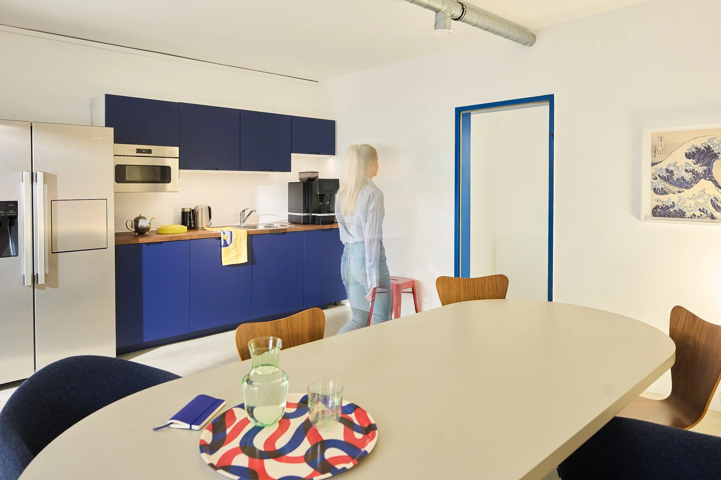 Küchendesign mit cobaltblauen Fronten und Edelstahldetails, großer grauer ovaler Tisch mit Holz- und Polsterstühlen, Hokusai Kunstdruck