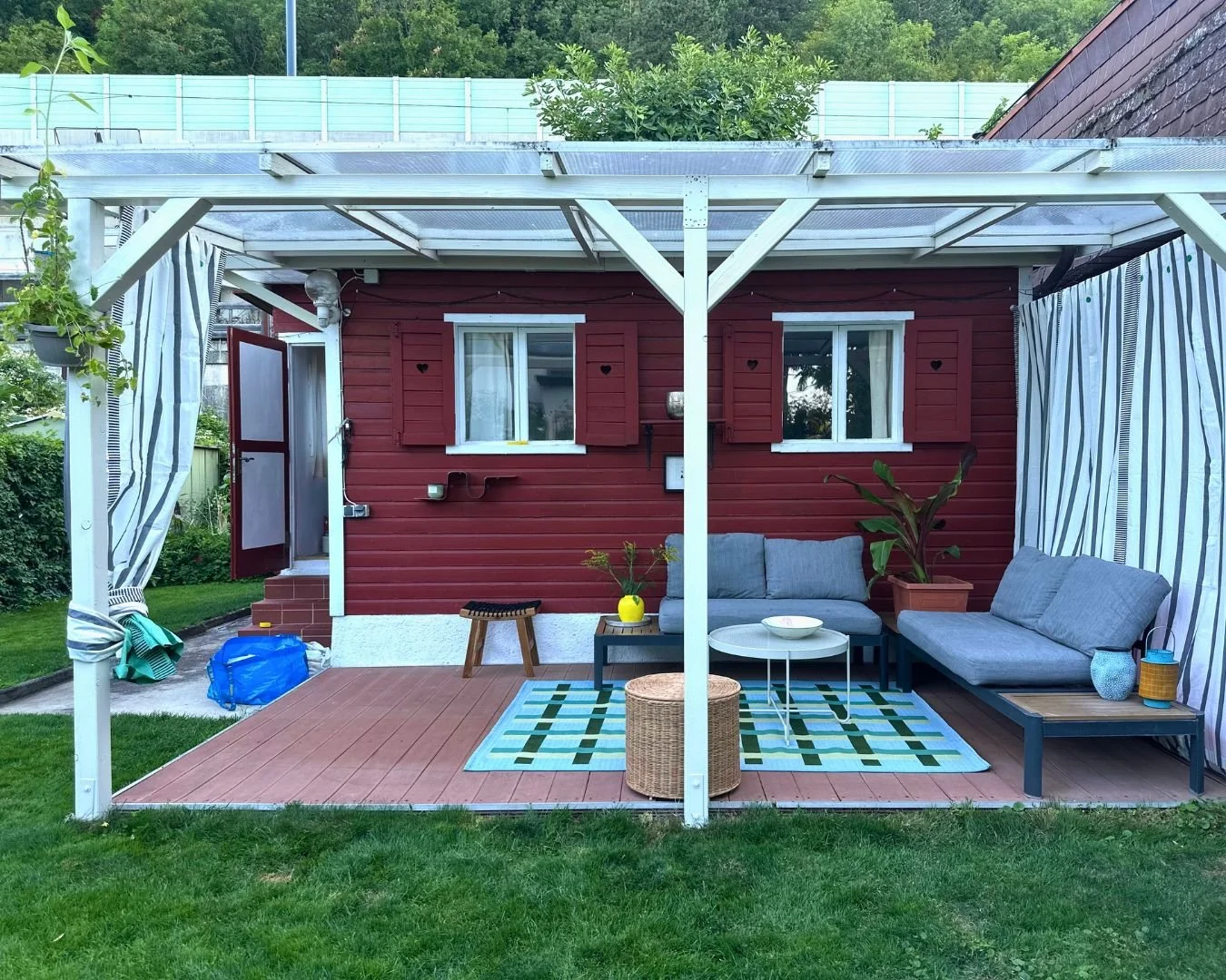 Gemütliche Terrasse mit einer Holzwand in Schwedenrot und gestreiften Vorhängen, geflochtener Hocker, Holzhocker. Geometrische Muster, Outdoorteppich, Outdoortisch, Outdoorgestaltung