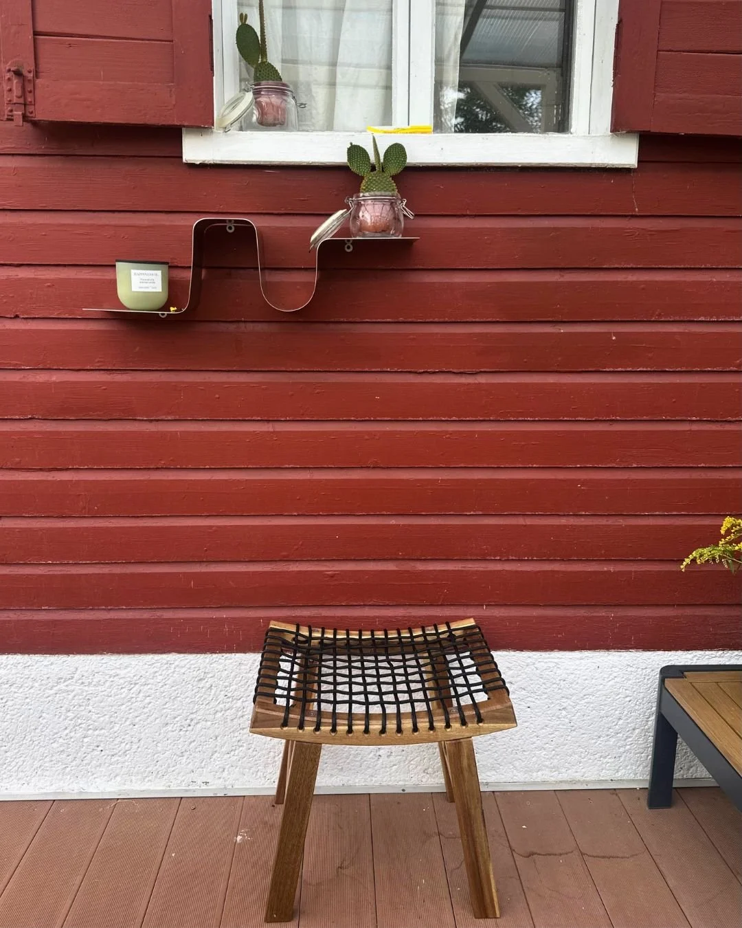 Detailaufnahme einer Terrasse, Holzwand in Schwedenrot, Holzhocker mit geflochtener Sitzfläche. Organisches Metallregal, Kakteen.
