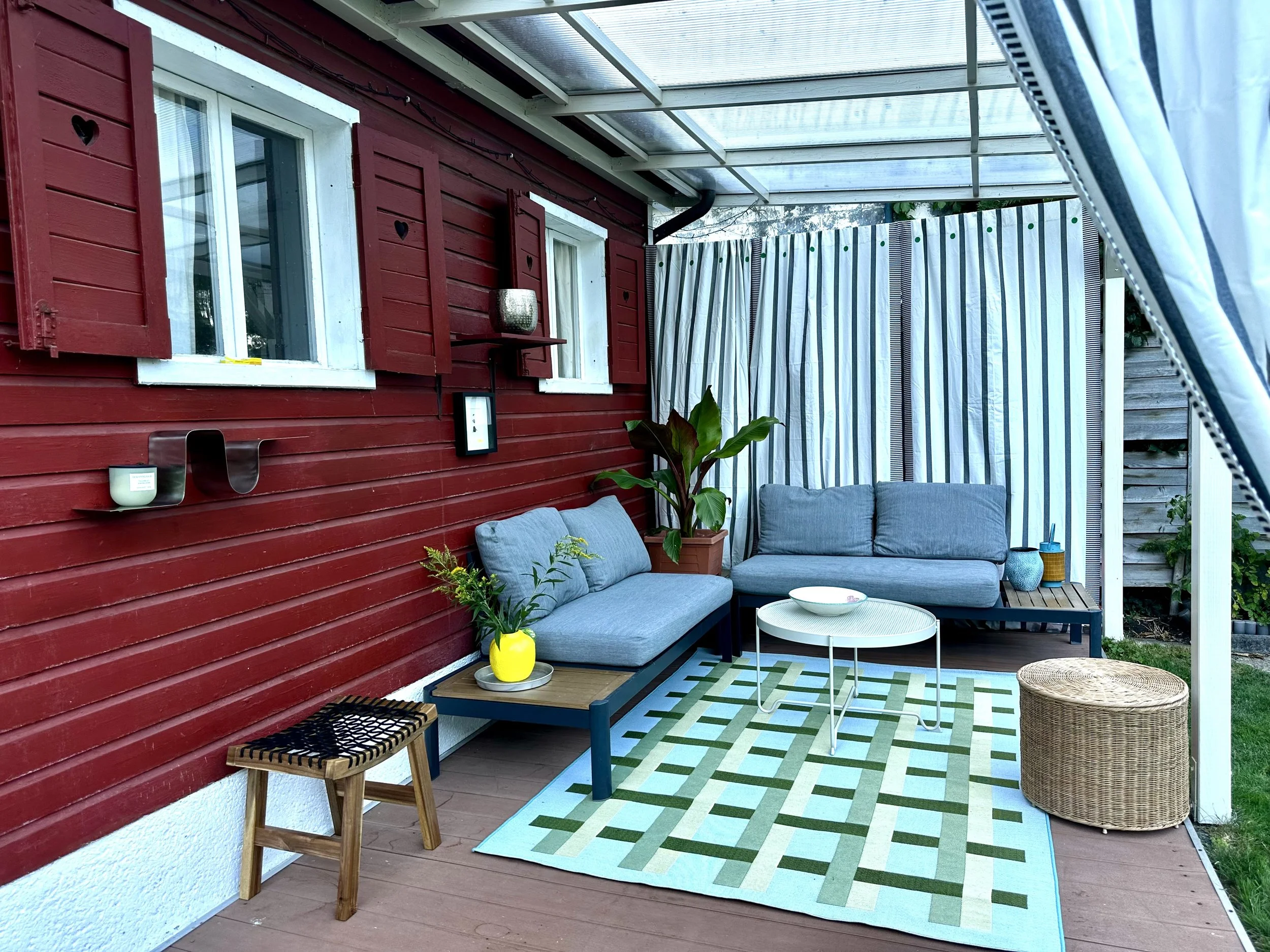 Gemütliche Terrasse mit einer Holzwand in Schwedenrot und gestreiften Vorhängen, geflochtener Hocker, Holzhocker. Geometrische Muster, Outdoorteppich, Outdoortisch, Outdoorgestaltung
