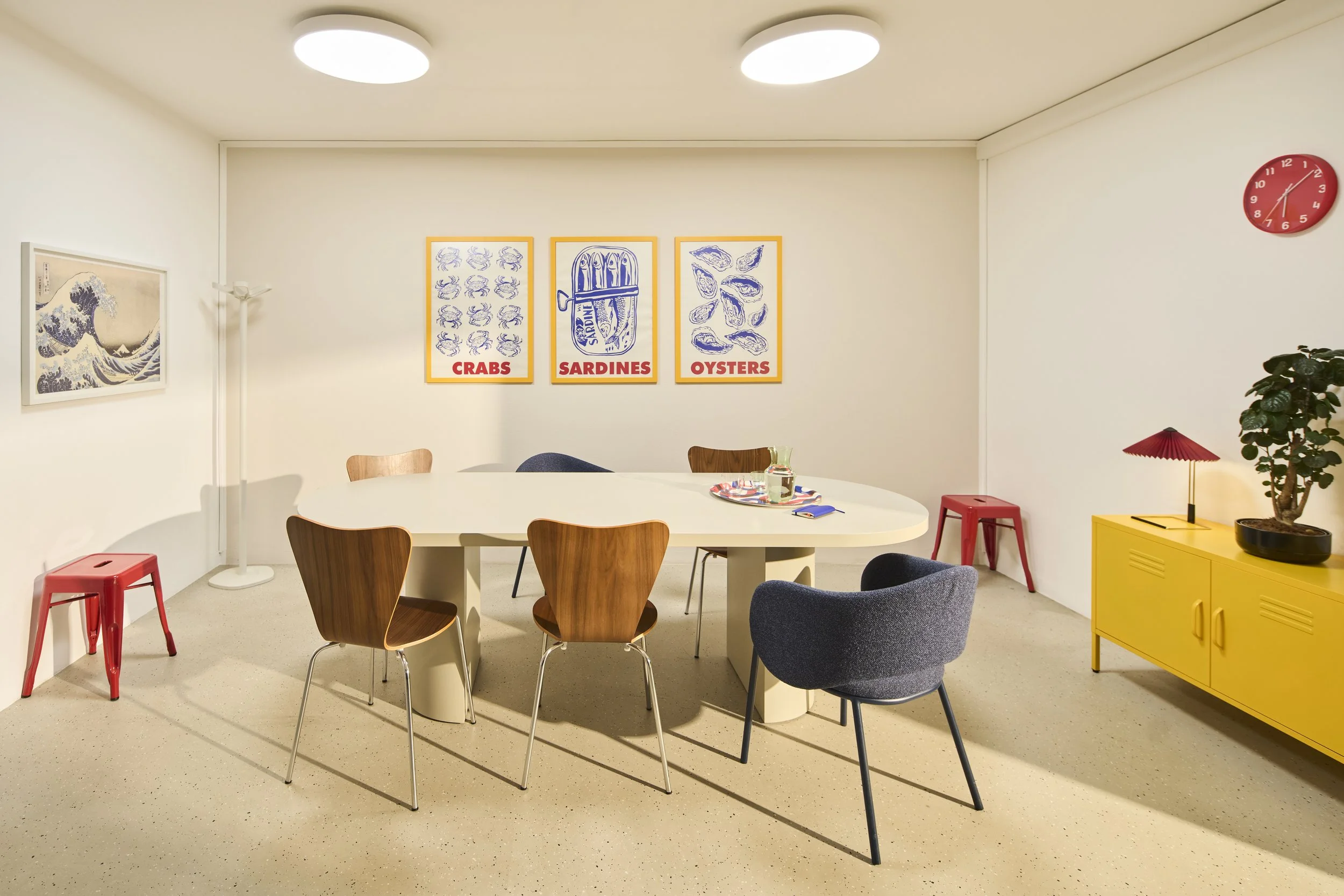 Farbenfrohe Teeküche und Aufenthaltsraum mit großem ovalem Tisch, um den herum Holz- und Polsterstühle stehen, einem gelben Metallschrank mit einer Pflanze und kleinen roten Hay-Leuchte, sowie drei Kunstdrucken an der Wand und roten Metallhockern.