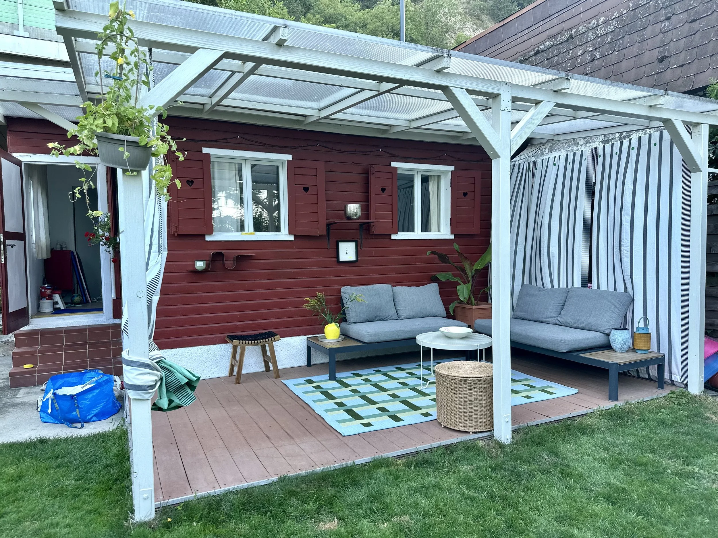Gemütliche Terrasse mit einer Holzwand in Schwedenrot und gestreiften Vorhängen, geflochtener Hocker, Holzhocker. Geometrische Muster, Outdoorteppich, Outdoortisch, Outdoorgestaltung