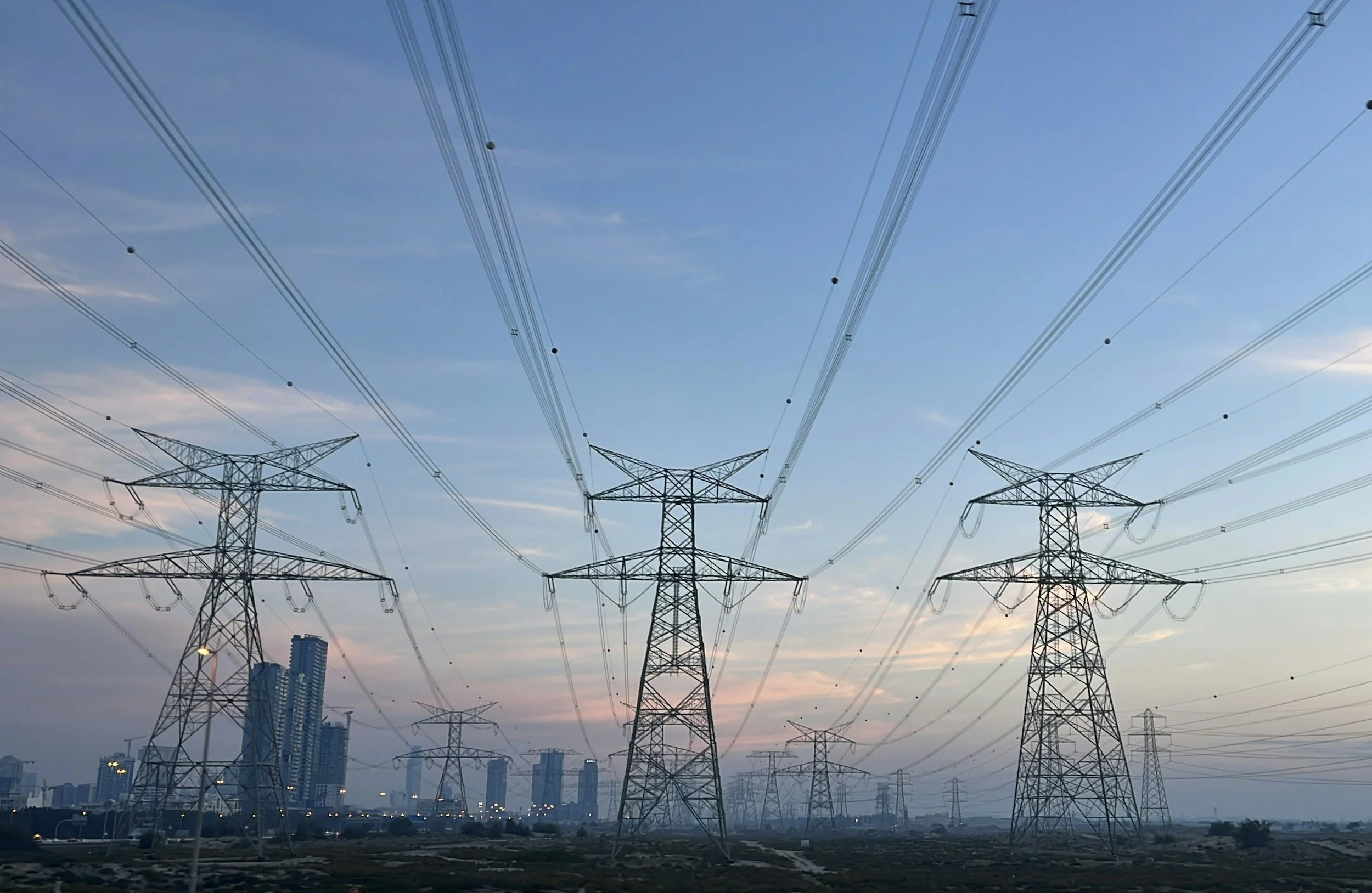 High-voltage power lines and pylons stretch across a cityscape during sunset, with tall buildings in the background.