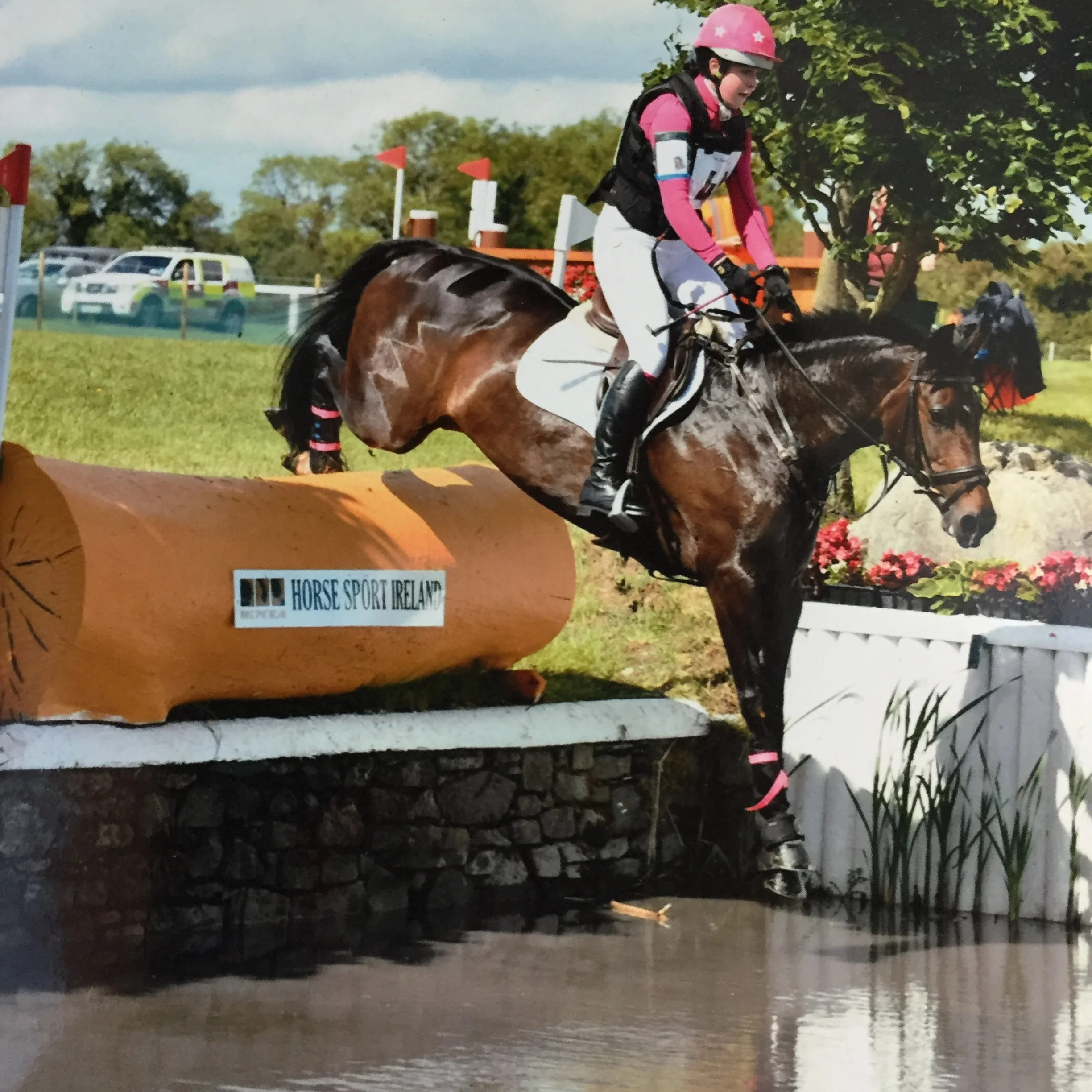 A young girl riding a horse during a horse riding event outdoors, with cars and trees in the background.