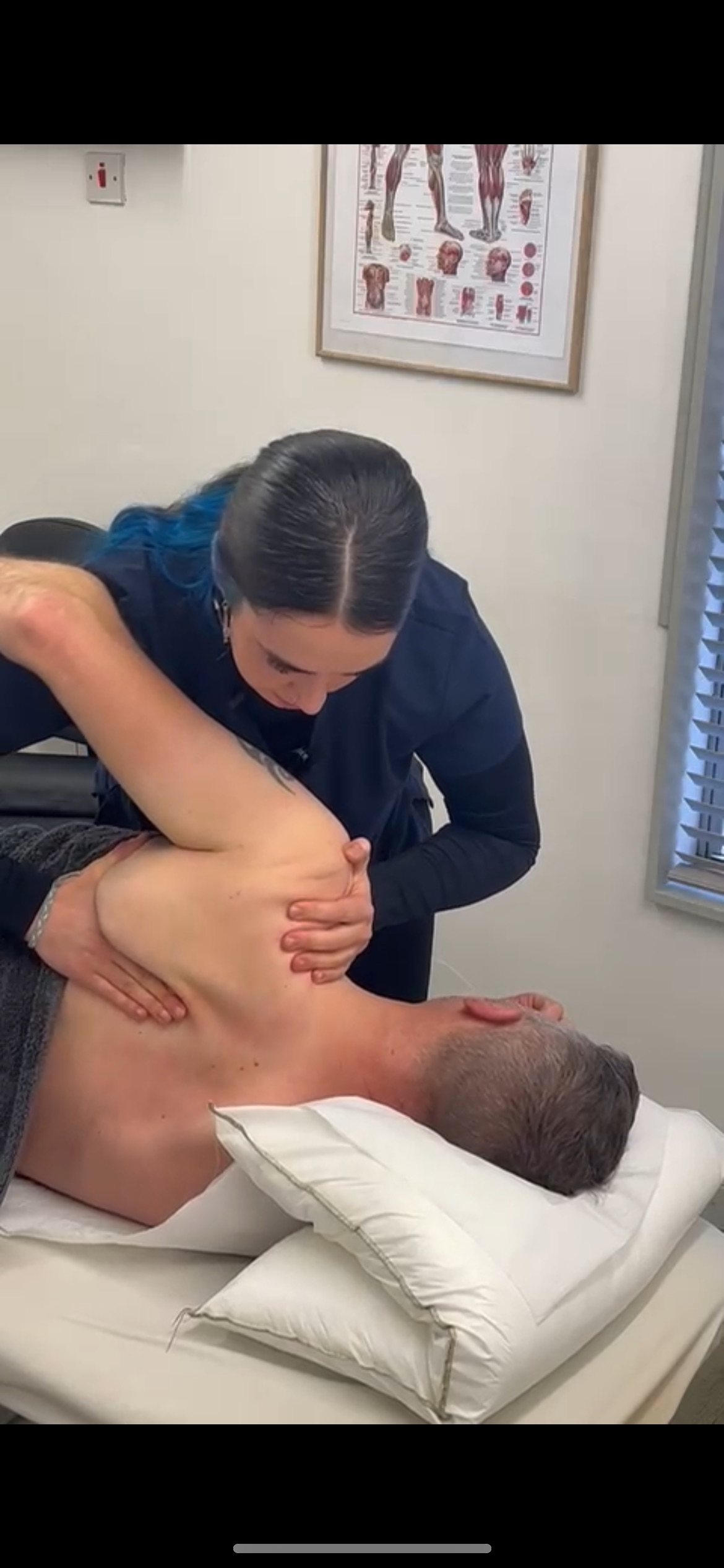 A person lying on a bed receiving a chiropractic adjustment from a healthcare professional in a clinic, with medical chart on the wall.