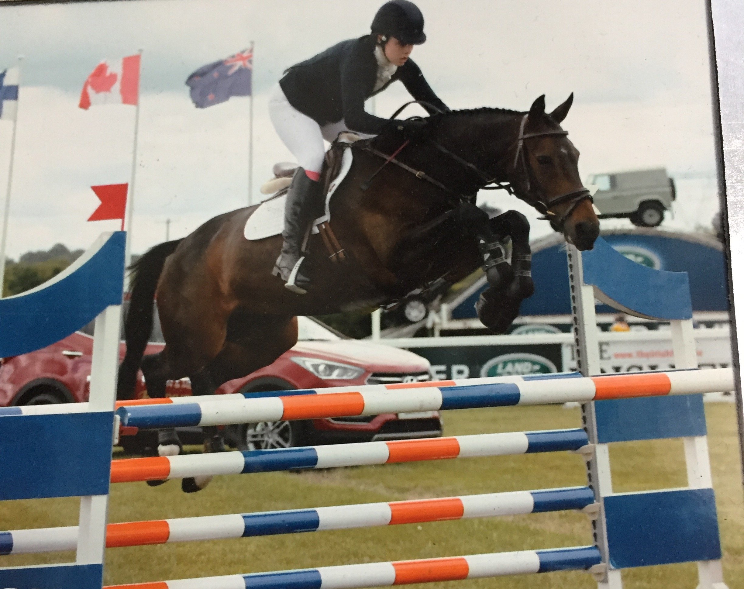 A female equestrian riding a brown horse with a white marking, jumping over an obstacle in a show jumping competition. She is wearing a black helmet and riding attire. There are flags in the background, including the Australian flag, along with cars parked and a cloudy sky.