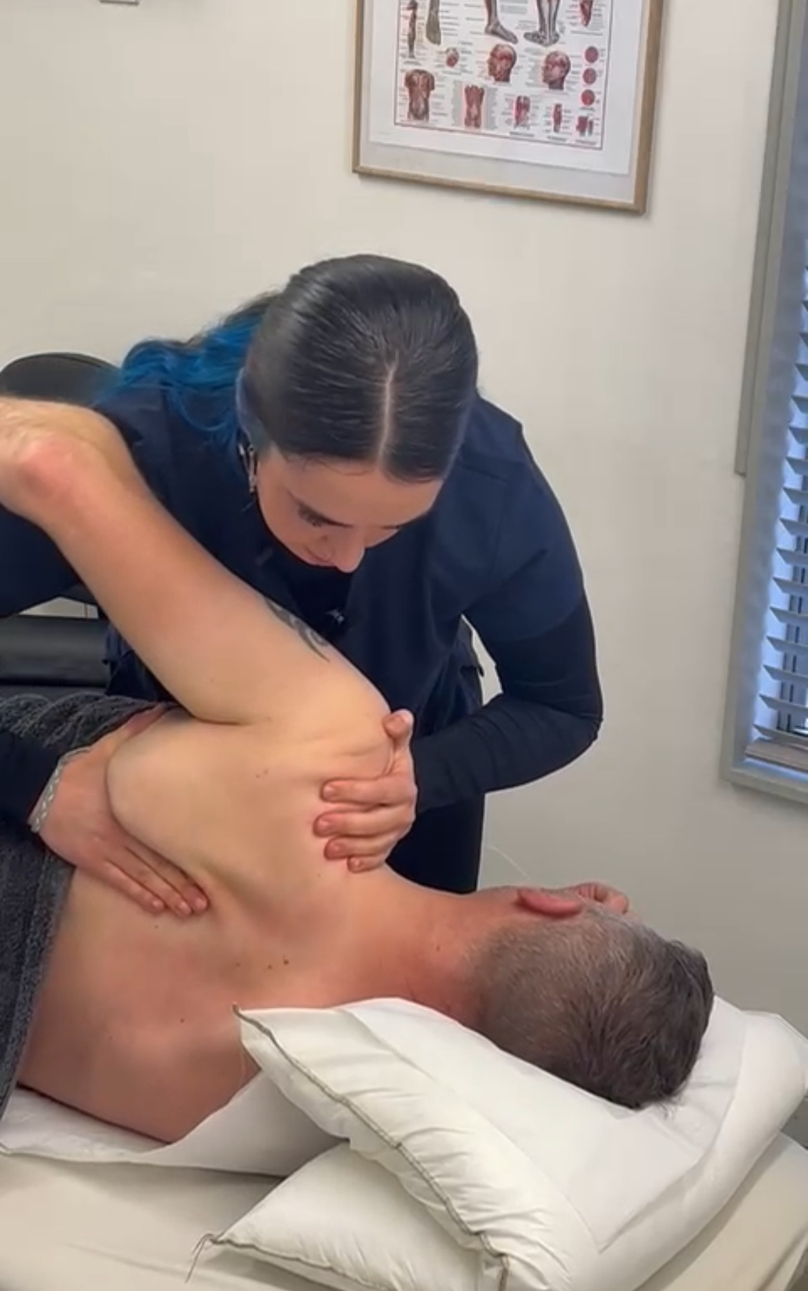 A person receiving a medical or chiropractic adjustment from a healthcare professional, lying on a bed with head on a pillow, in a clinical setting with a medical chart hanging on the wall.