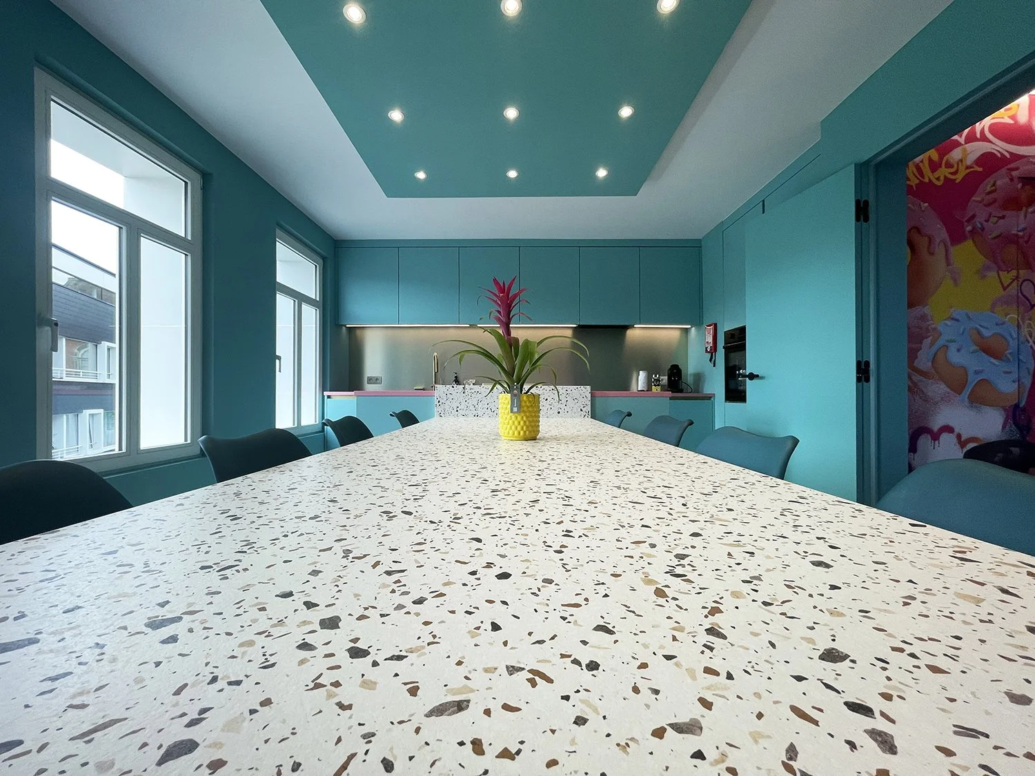 Salle de réunion moderne avec une grande table en terrazzo, un vase avec une plante rose au centre, murs turquoise, fenêtres larges et cuisine en arrière-plan.