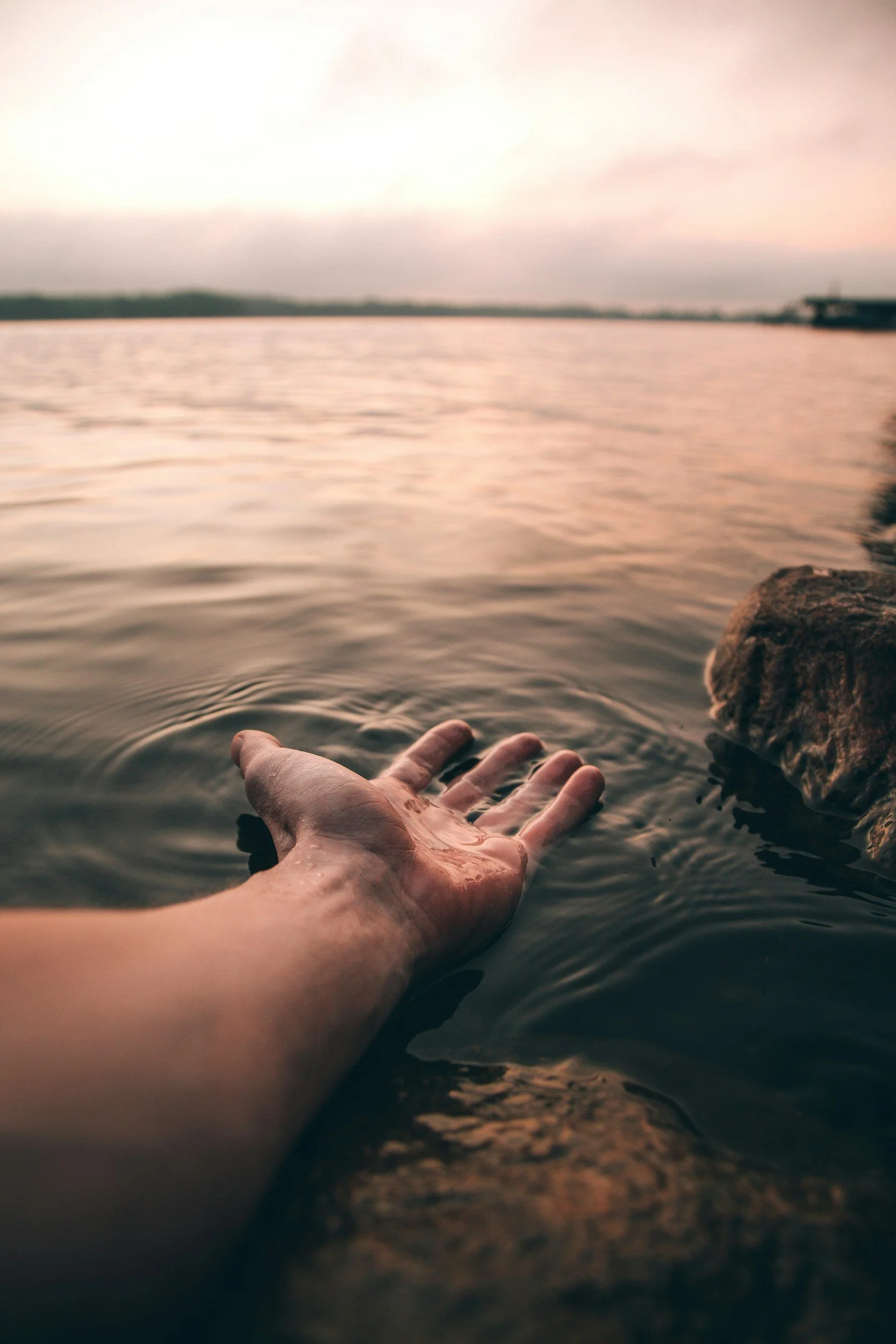 Close-up of a person's hand resting on the edge of a body of water, with rocks and a distant shoreline in the background during sunset.