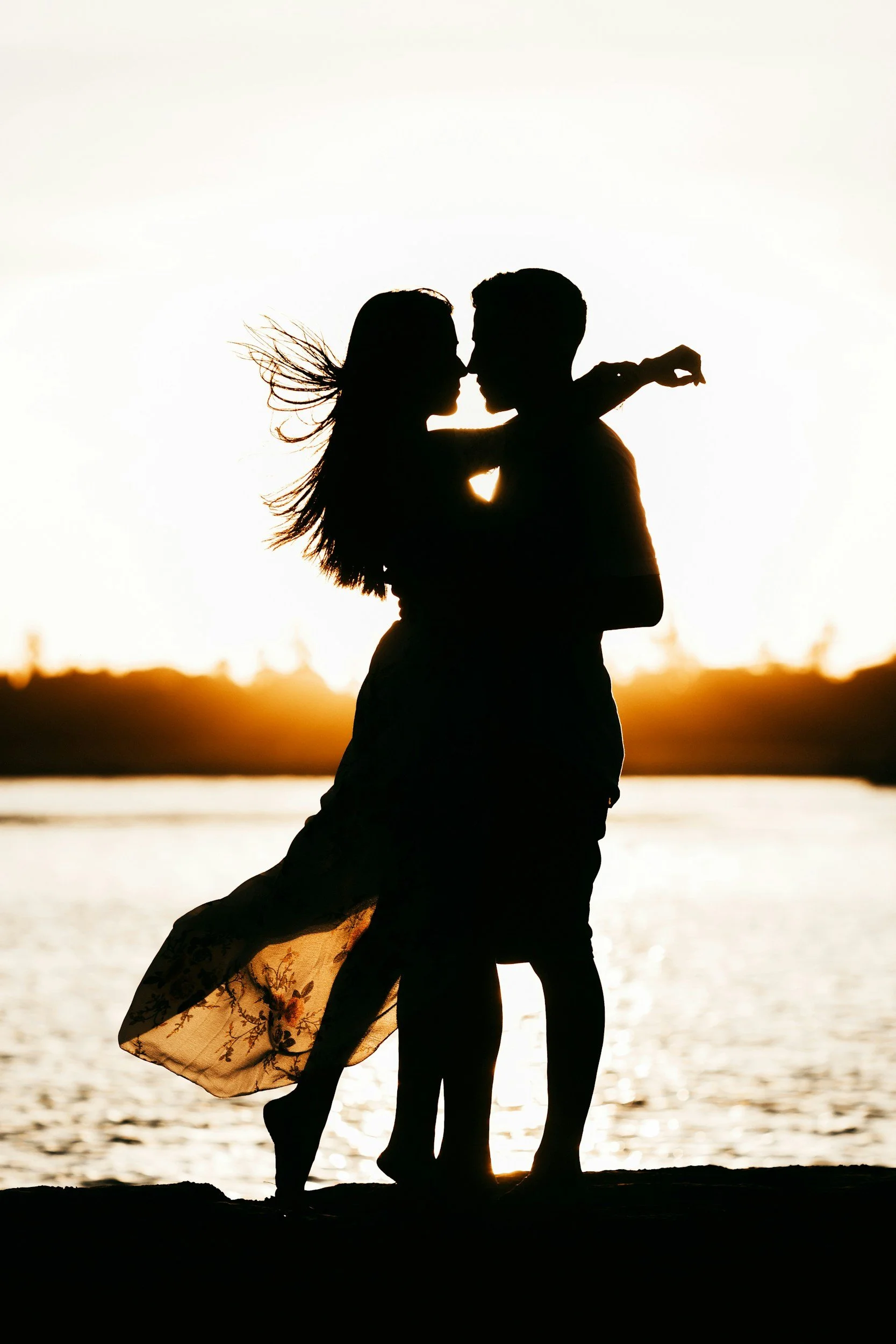 Silhouette of a couple dancing on the beach at sunset, with water and a distant horizon in the background.