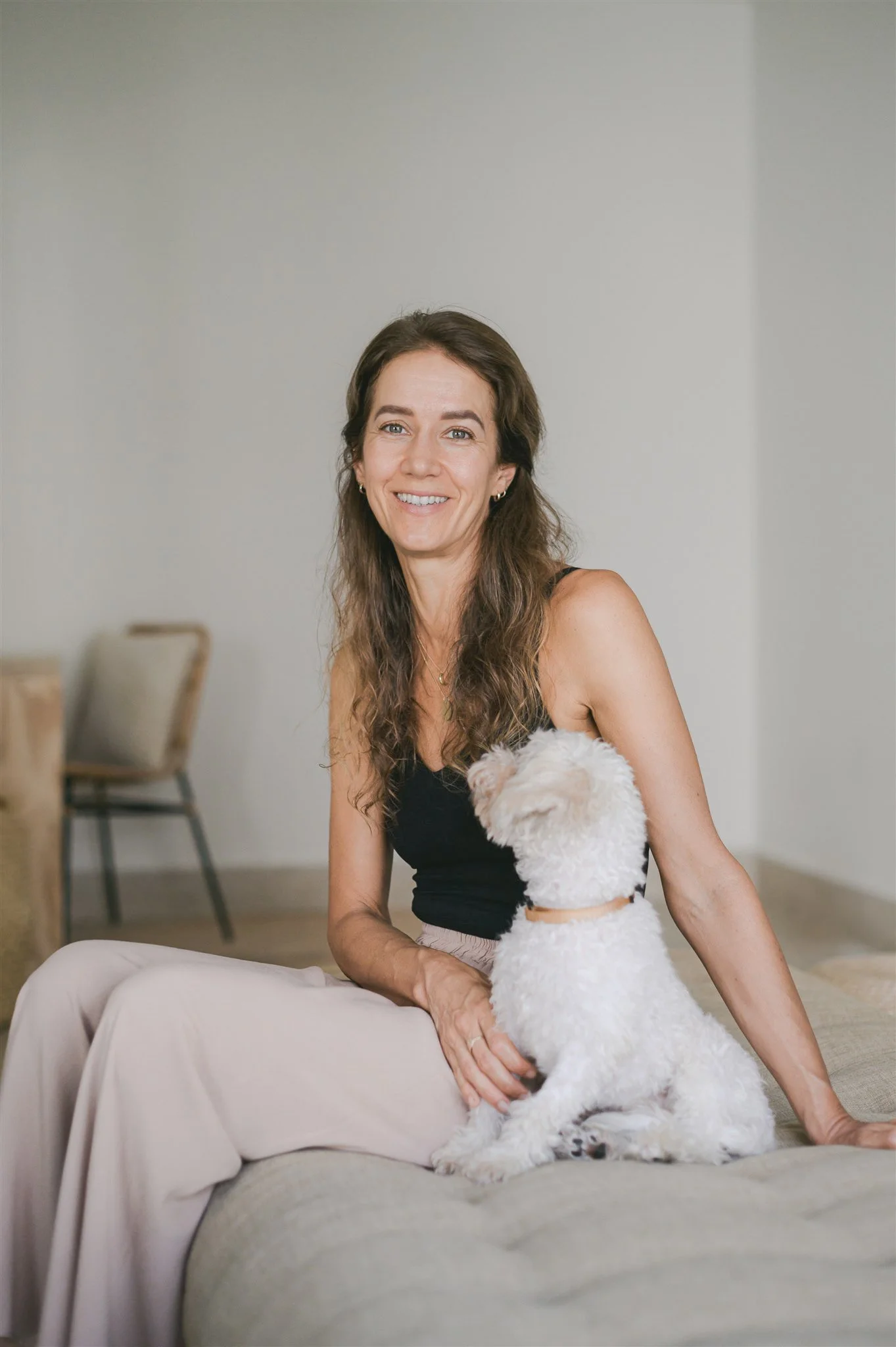 A woman with long wavy brown hair, wearing a black tank top and beige pants, smiling while sitting on a couch. She is holding a white fluffy dog, possibly a Bichon Frise, who is looking up at her. The background shows a plain light-colored wall and dining chairs.