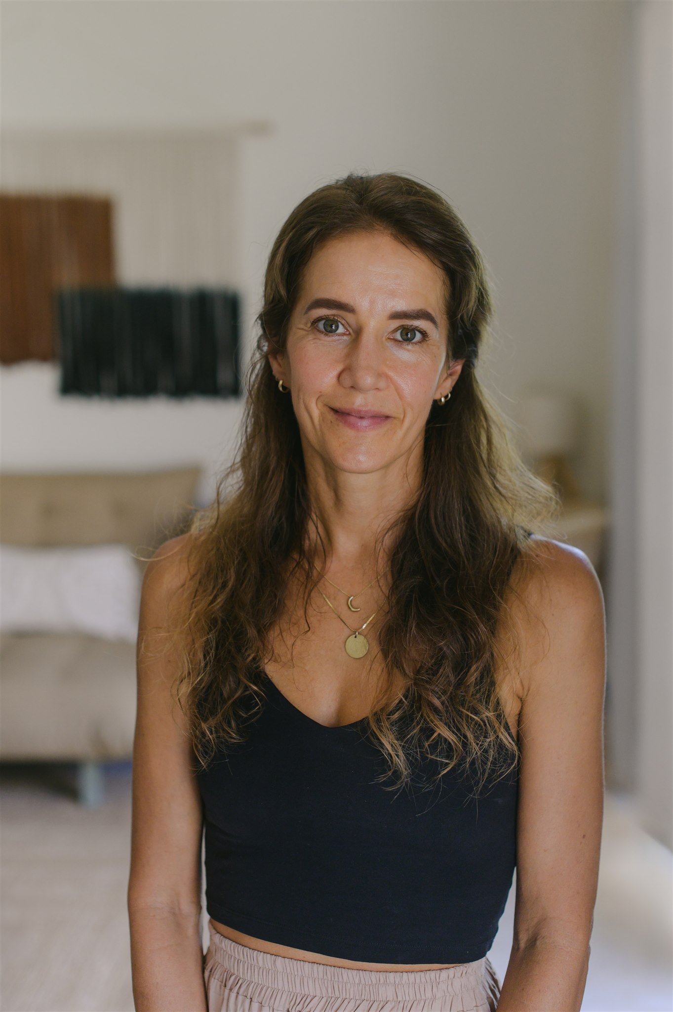 A woman with long wavy brown hair, wearing a black tank top, layered necklaces, and beige pants, standing in a neutral-toned room with a bed and wall decor in the background.