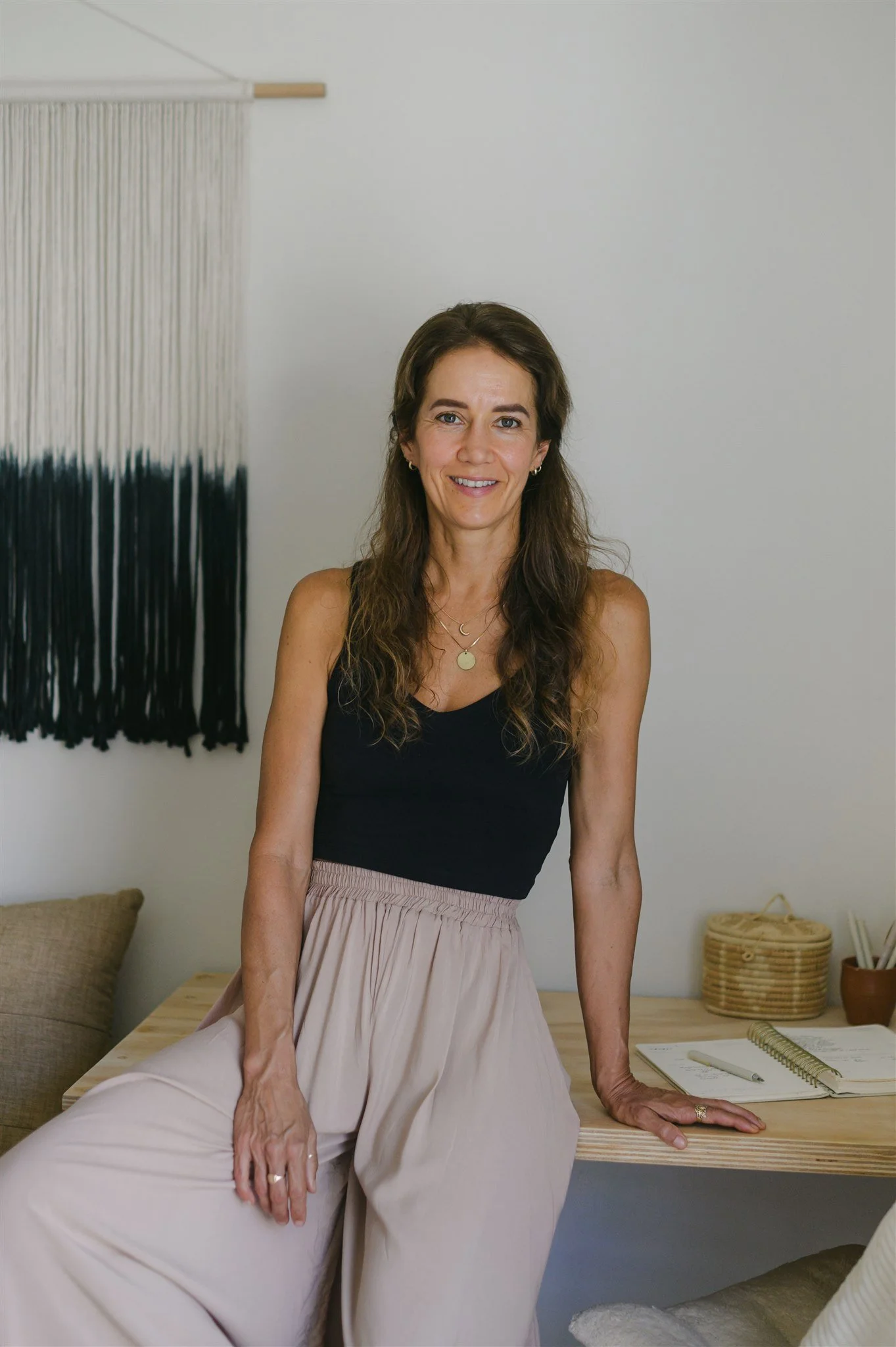 A woman with long brown hair, wearing a black sleeveless top and a beige skirt, sitting on a wooden table in a cozy room with neutral-colored decor, a woven wall hanging, and a notebook with a pen nearby.