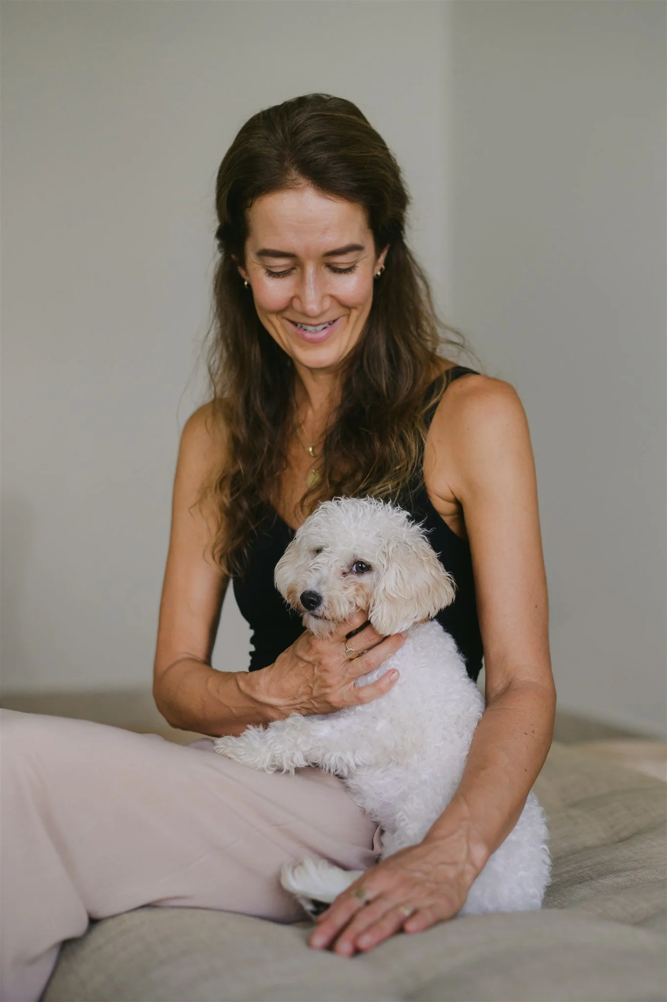 A woman with long brown hair, wearing a black sleeveless top, holding a white fluffy dog with one eye winking, smiling and looking down. The woman is sitting on a light-colored sofa with a gray background.