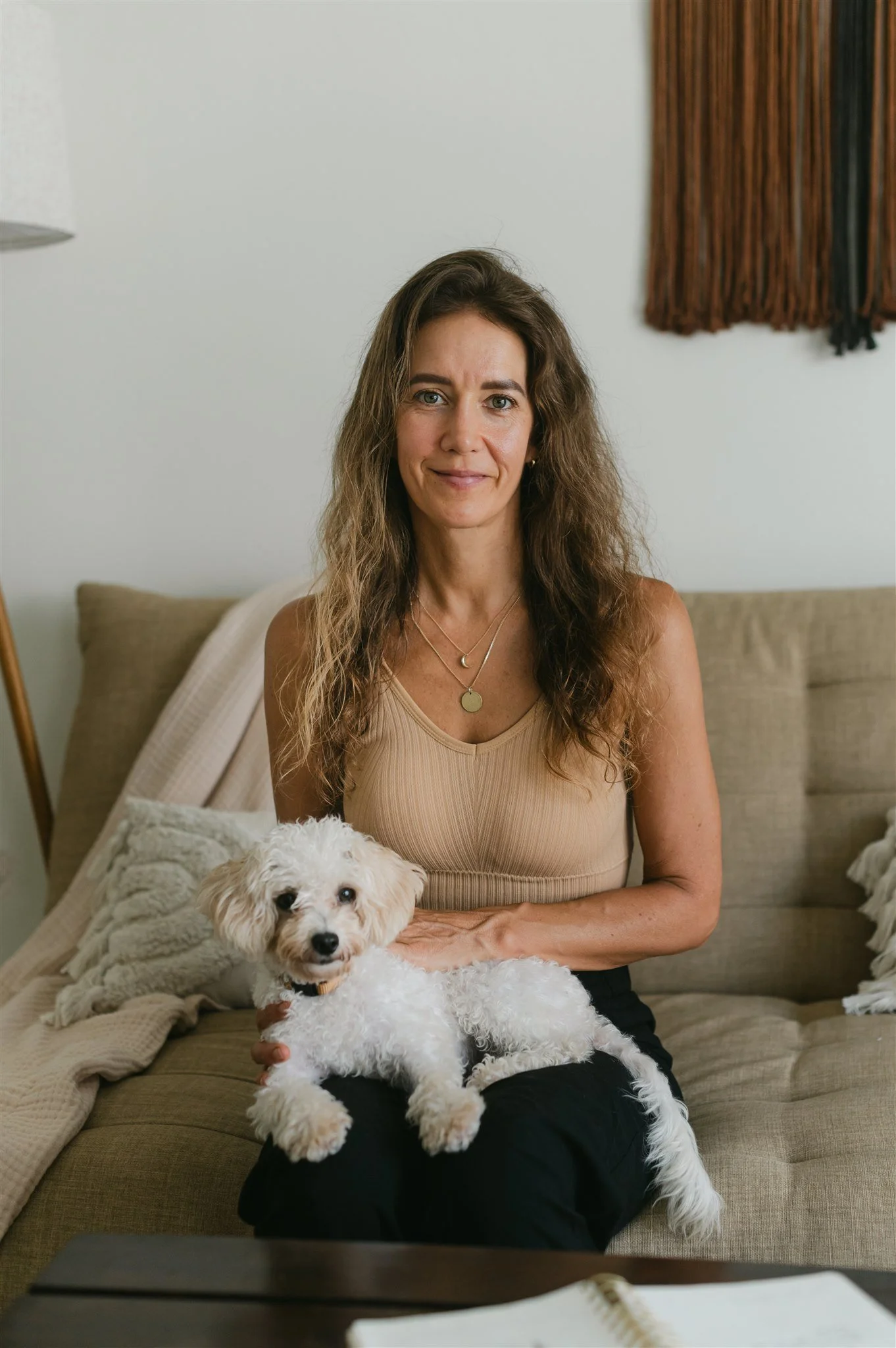 A woman with long curly hair sitting on a beige couch, holding a small white poodle dog. The woman is smiling and wearing a beige tank top with layered necklaces. There are blankets on the couch and a wall with a brown textured wall hanging in the background.