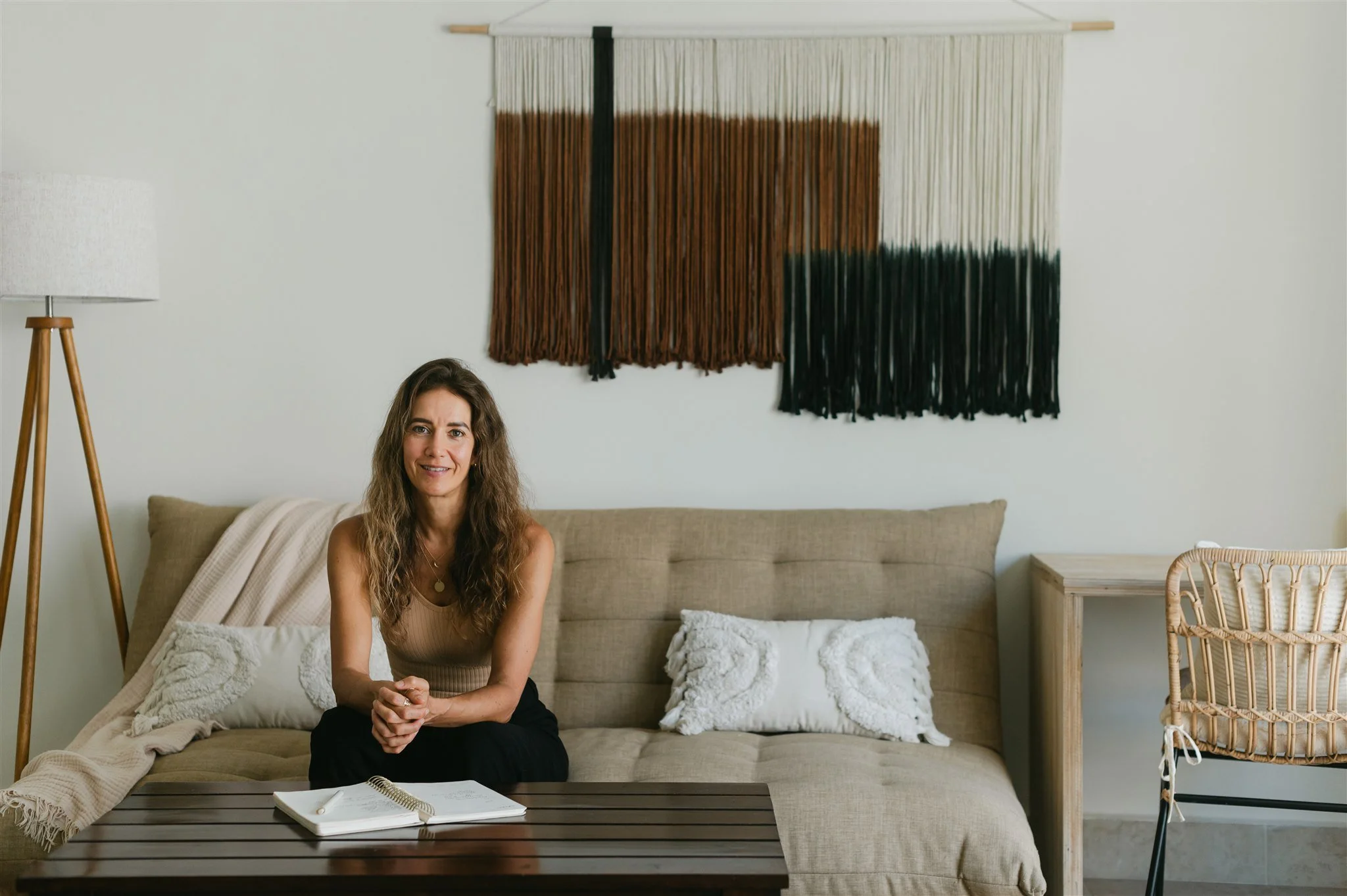 A woman with wavy brown hair sitting on a beige sofa with white pillows in a modern, minimalistic living room. Behind her, there is a wall with hanging art that features vertical strands in shades of brown, cream, and black. To her right, there is a side table and a rattan chair; a wooden floor lamp is on her left.