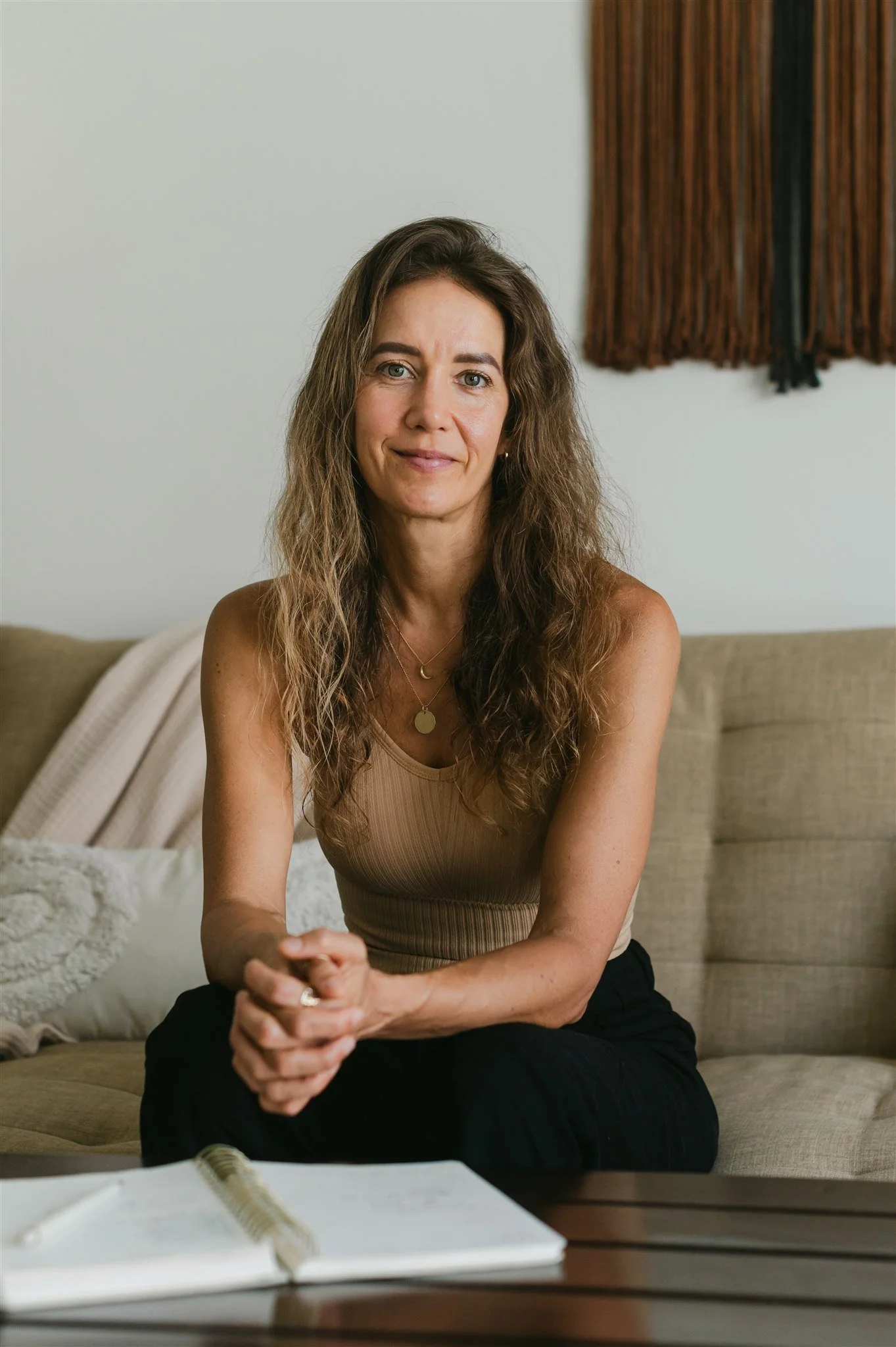 A woman with long wavy hair, wearing a beige tank top, sitting on a beige couch in a room with a white wall and a hanging brown textured art piece, holding her hands together, with an open notebook on the table in front of her.