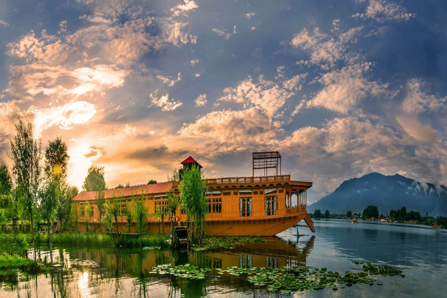 A floating wooden house on a calm lake with lily pads, green trees, mountains in the background, and a colorful sunset sky.