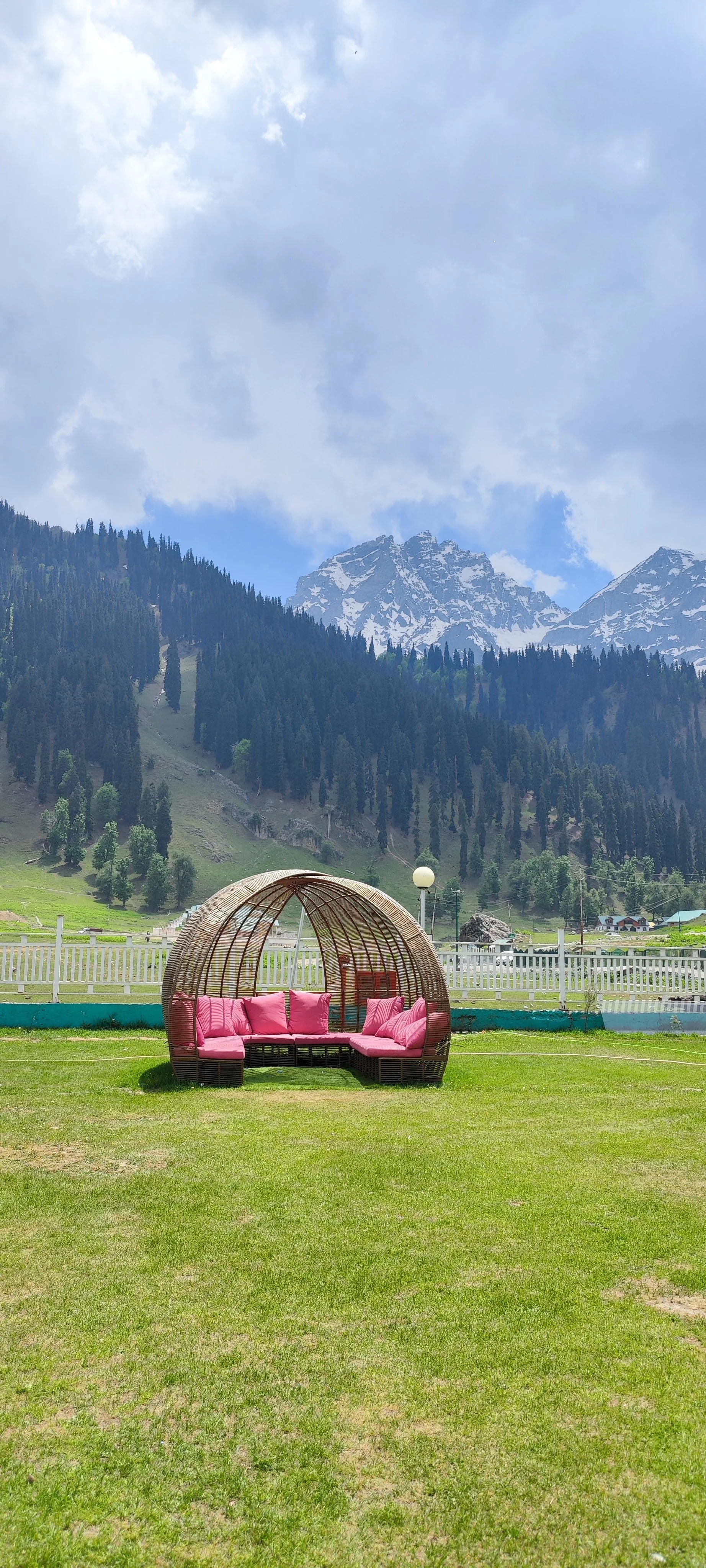 A wicker outdoor sofa with pink cushions on a grassy lawn, with snow-capped mountains and a cloudy sky in the background.