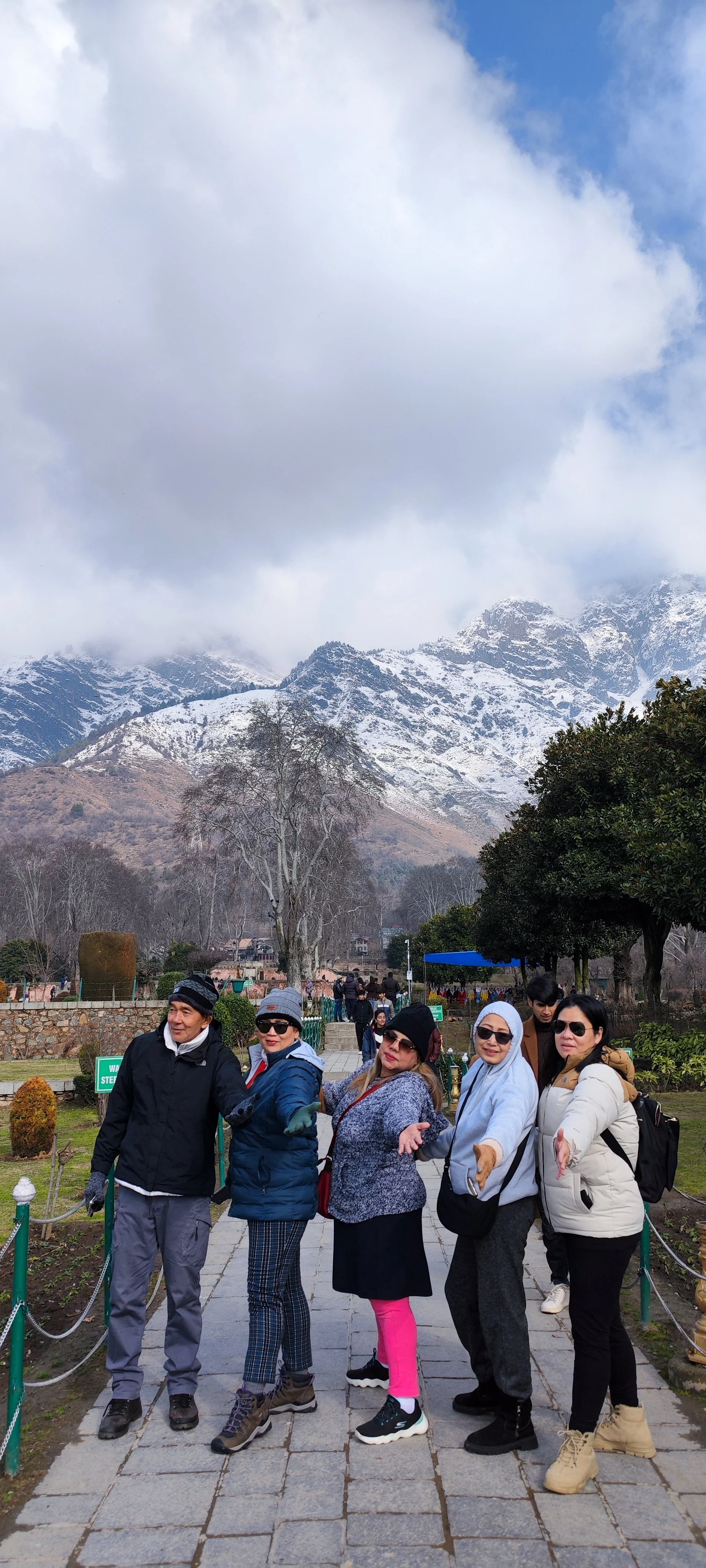 Group of five people wearing winter clothing, standing on a paved pathway in a park with mountain scenery, snow-capped peaks, and partly cloudy sky in the background.
