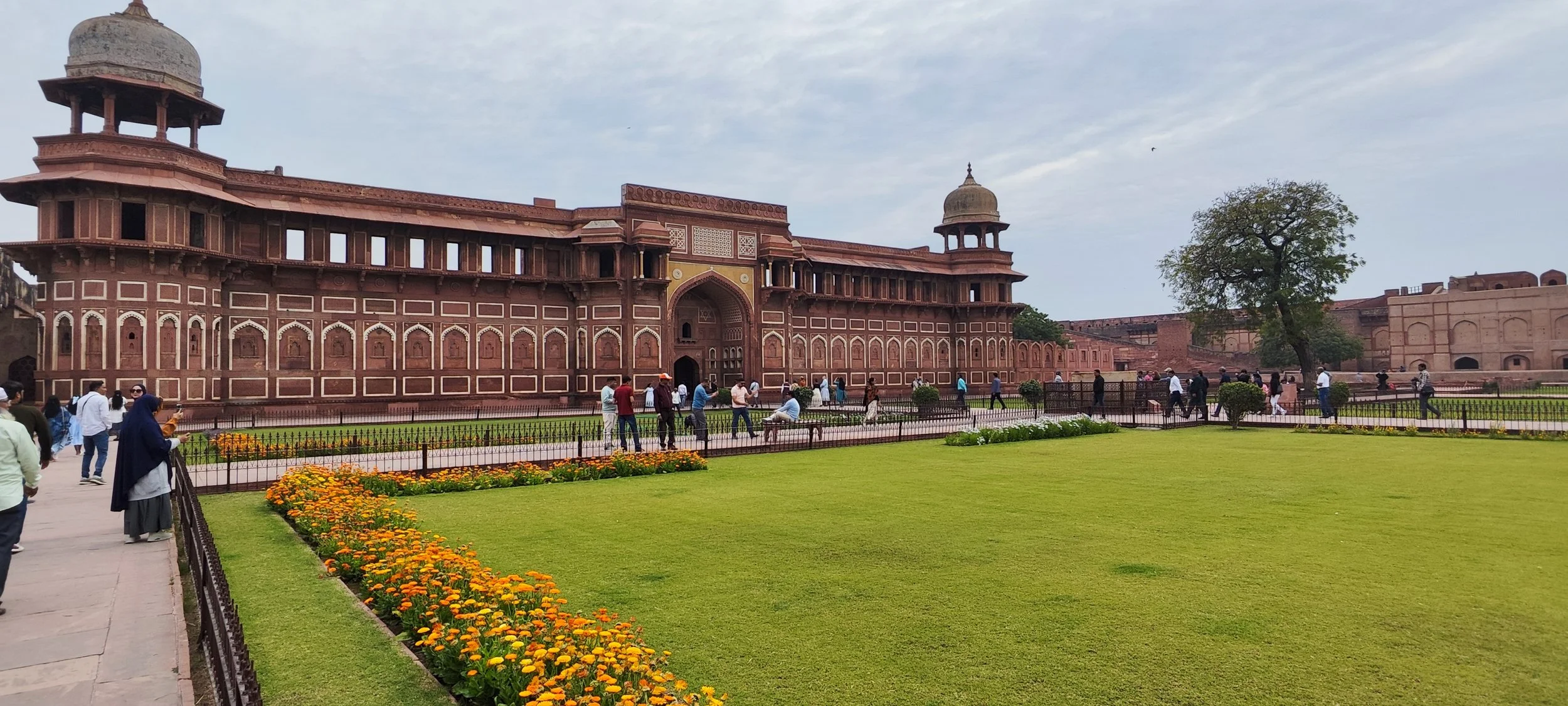 Historic red sandstone building with intricate carvings, surrounded by a garden with green grass, orange flowers, and a few trees, with tourists walking around.