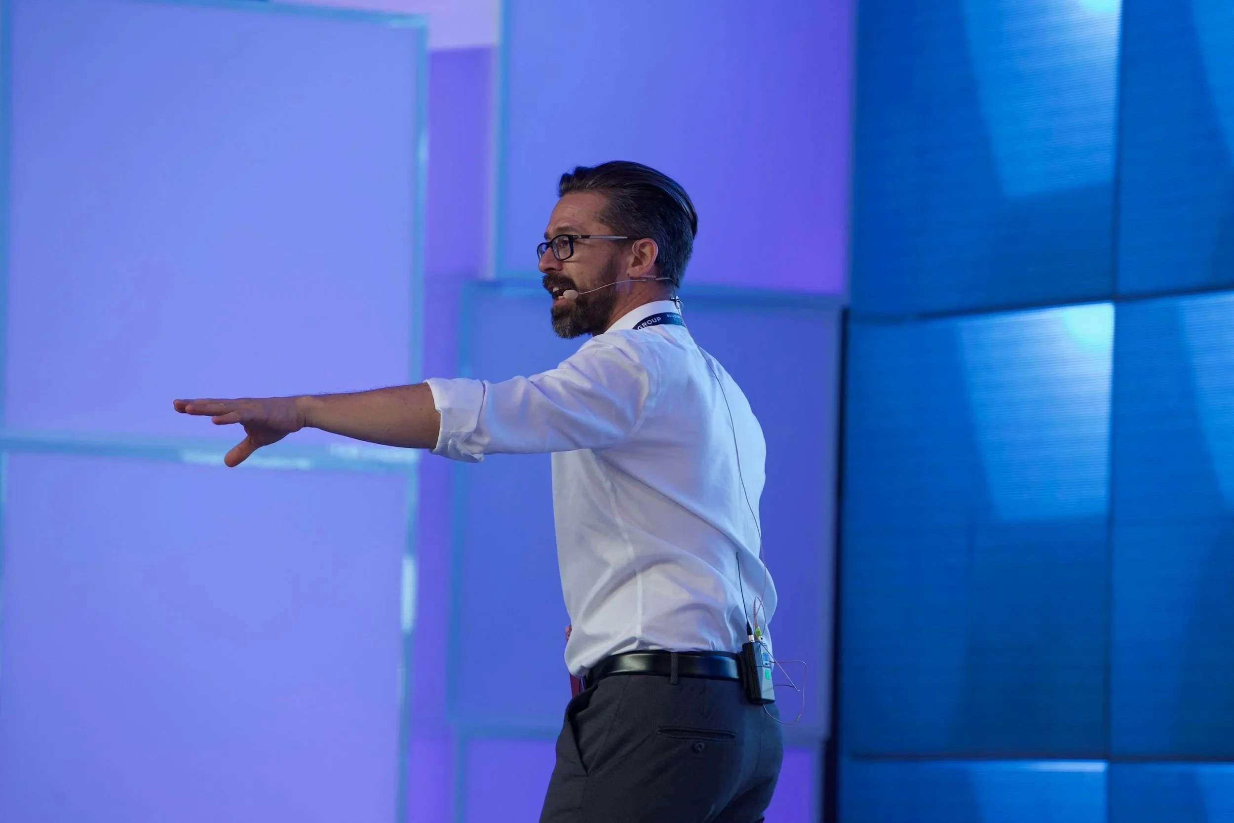 A man wearing glasses and a white shirt giving a presentation on stage, with blue and purple lighting in the background.