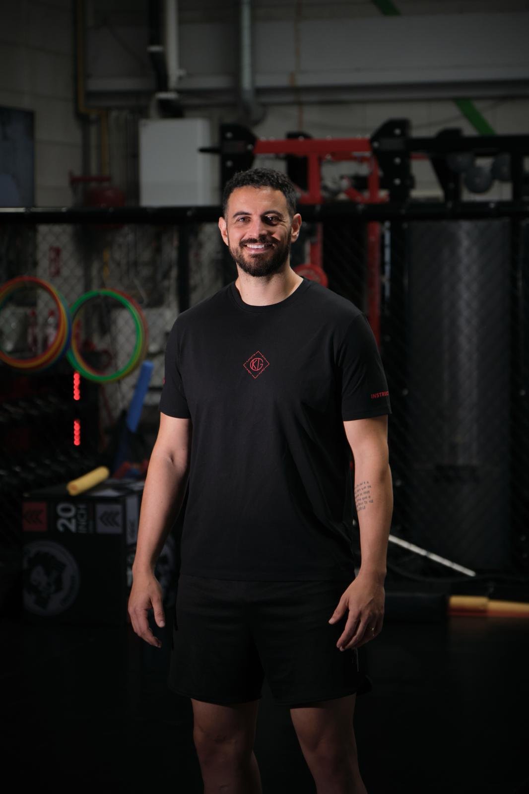 Man standing in a gym, smiling, wearing a black t-shirt and shorts, with gym equipment and black fencing in the background.