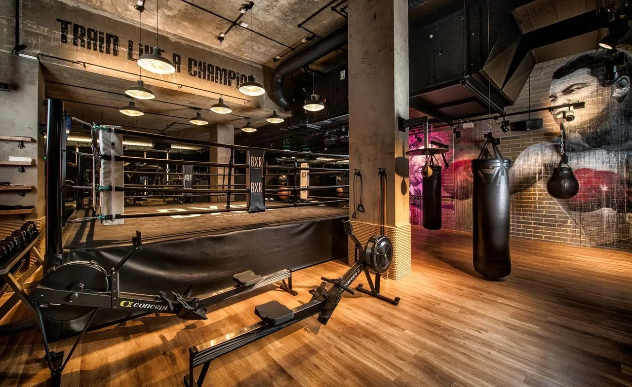 Empty boxing gym with a boxing ring, punching bags, and workout equipment on wooden floors and exposed brick walls with a mural of a boxer.