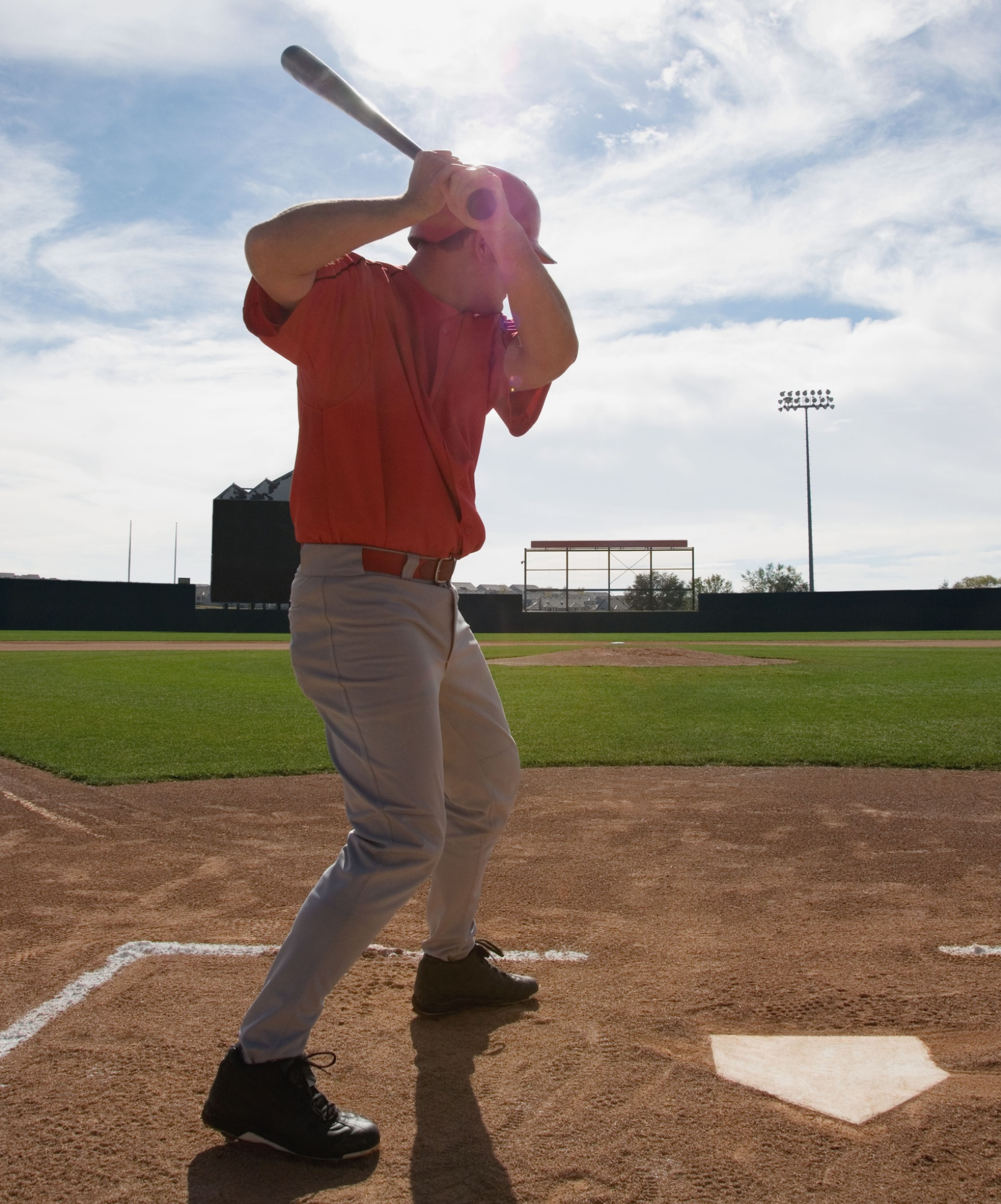 A baseball player in a red shirt and white pants is preparing to hit a pitched ball on a baseball field with a bright sky and stadium lights in the background.