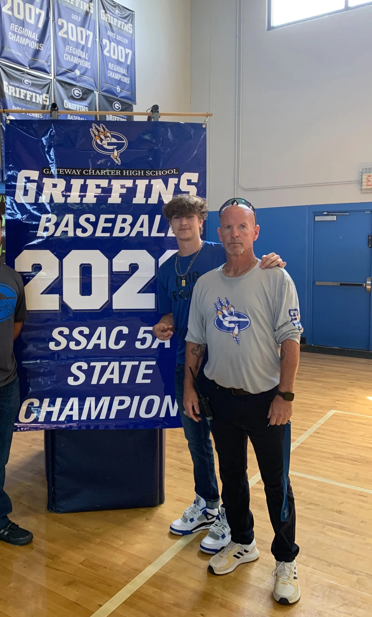 A man and a young man standing in a gymnasium, posing in front of a championship banner that reads 'Gateway Charter High School Griffins Baseball 2022 SSAC 5A State Champions.' The man has his arm around the young man and is wearing a gray Griffins sweatshirt, while the young man is in a blue Griffins shirt and sneakers. Both are looking at the camera.