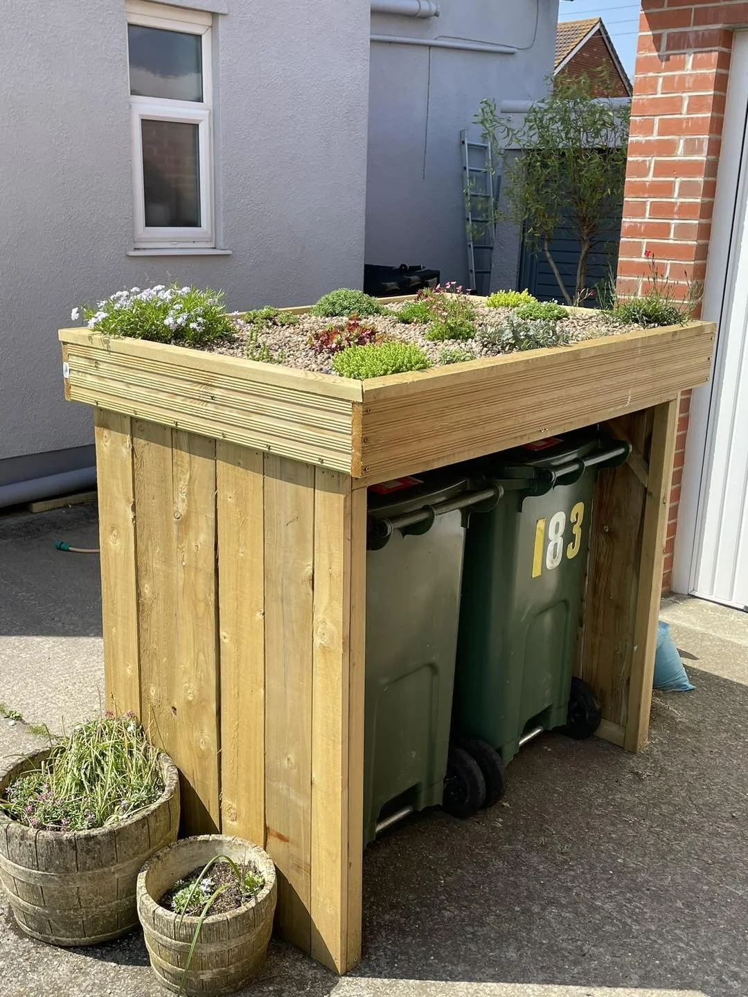 A wooden planter box containing various plants and flowers, placed above two green trash bins and two large flower pots with plants, situated outdoors next to a building and a brick wall.