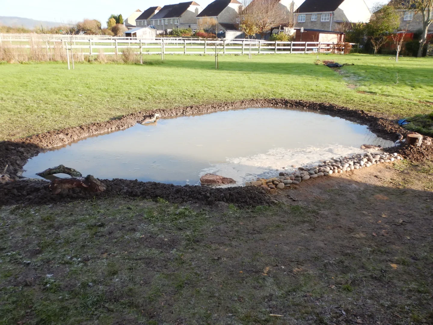 A small construction site with a dug-out pond bordered with rocks in a grassy backyard, with houses and trees in the background on a sunny day.