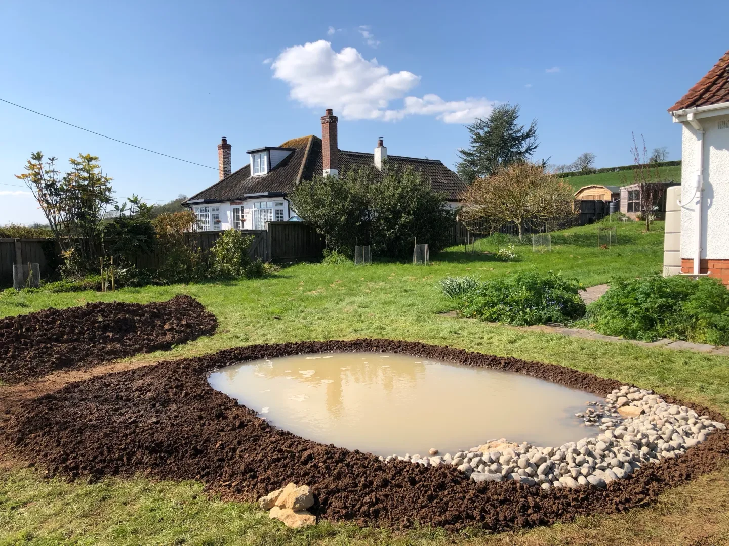 A backyard with a water-filled pond under construction, surrounded by dark soil, with a white house, garden, trees, and blue sky with clouds in the background.