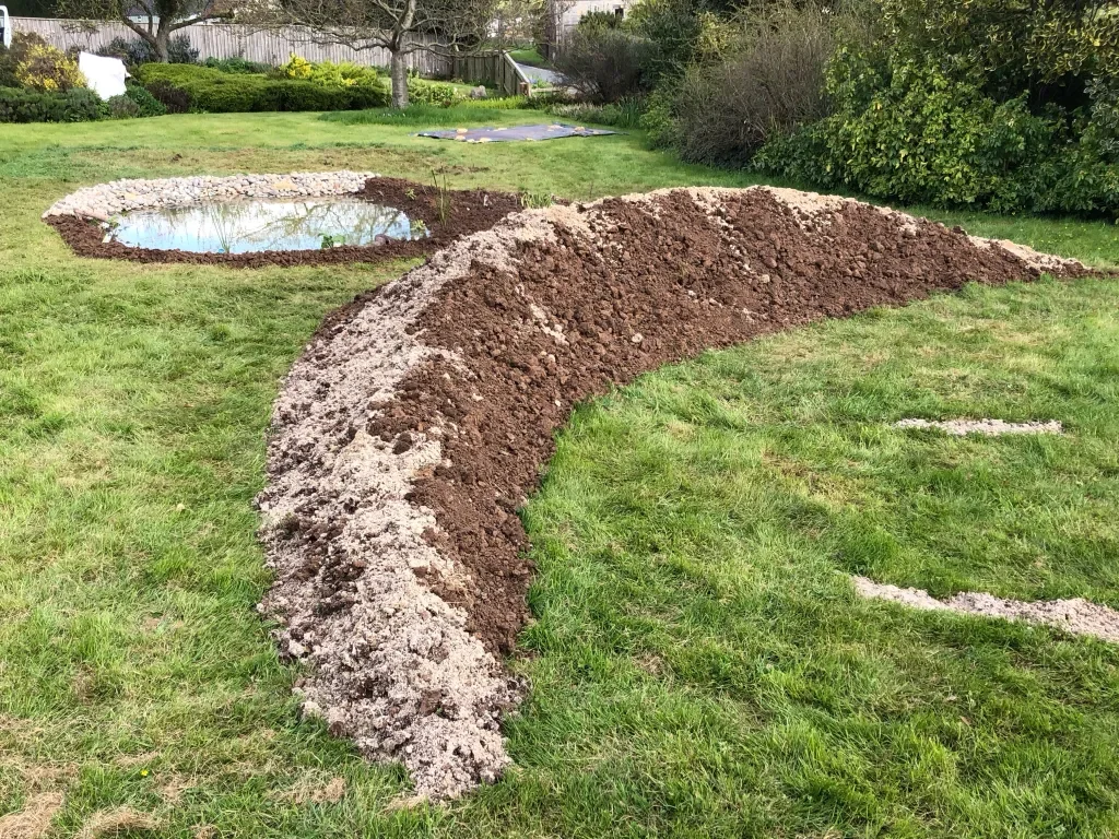A backyard with a pond and a large, arched trench of freshly dug soil.