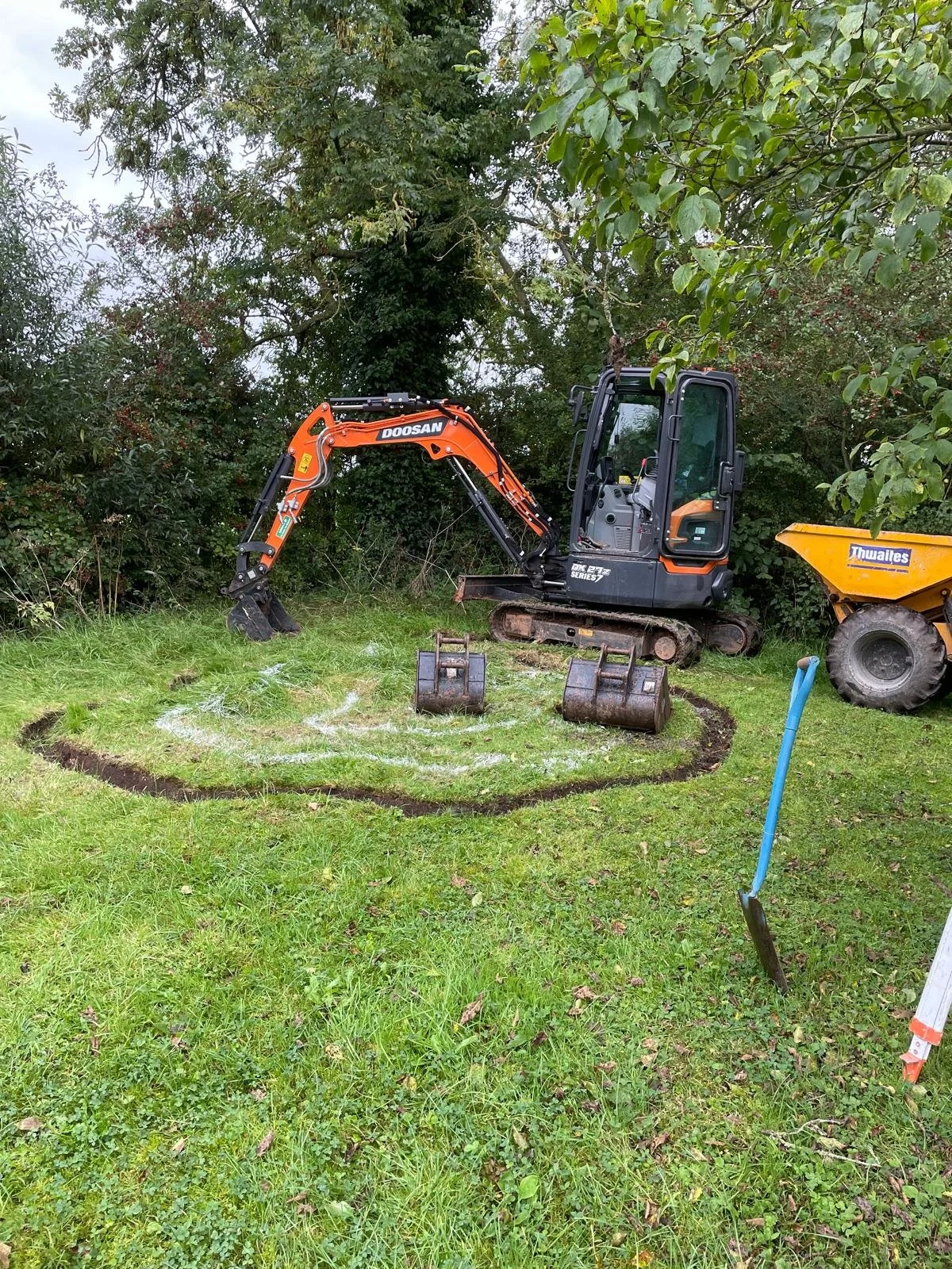 A small orange and black Doosan excavator digging in a grassy yard, surrounded by trees and bushes, with a blue shovel in the foreground and construction equipment nearby.