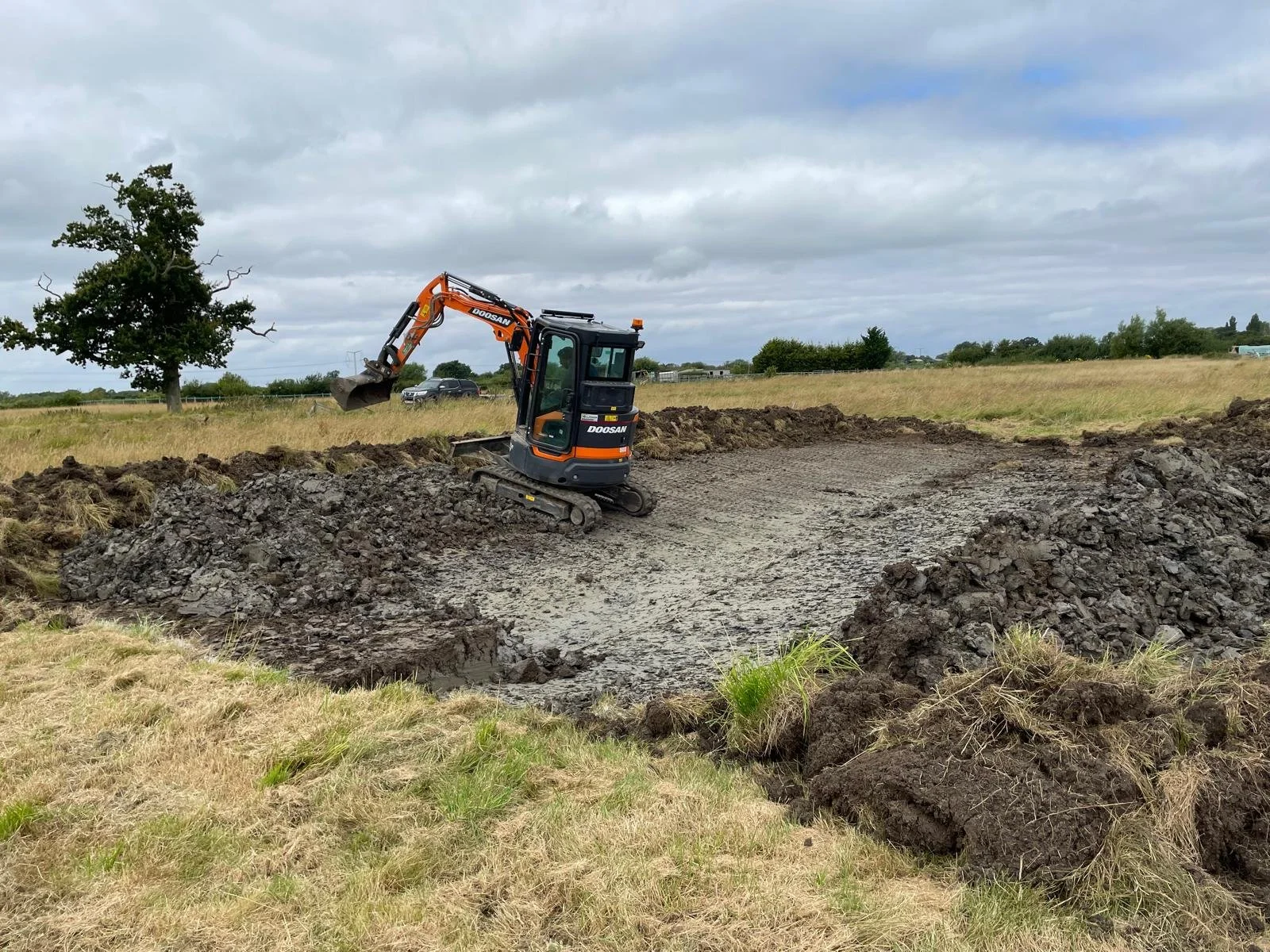 Small excavator digging a hole in a grassy field with a cloudy sky.