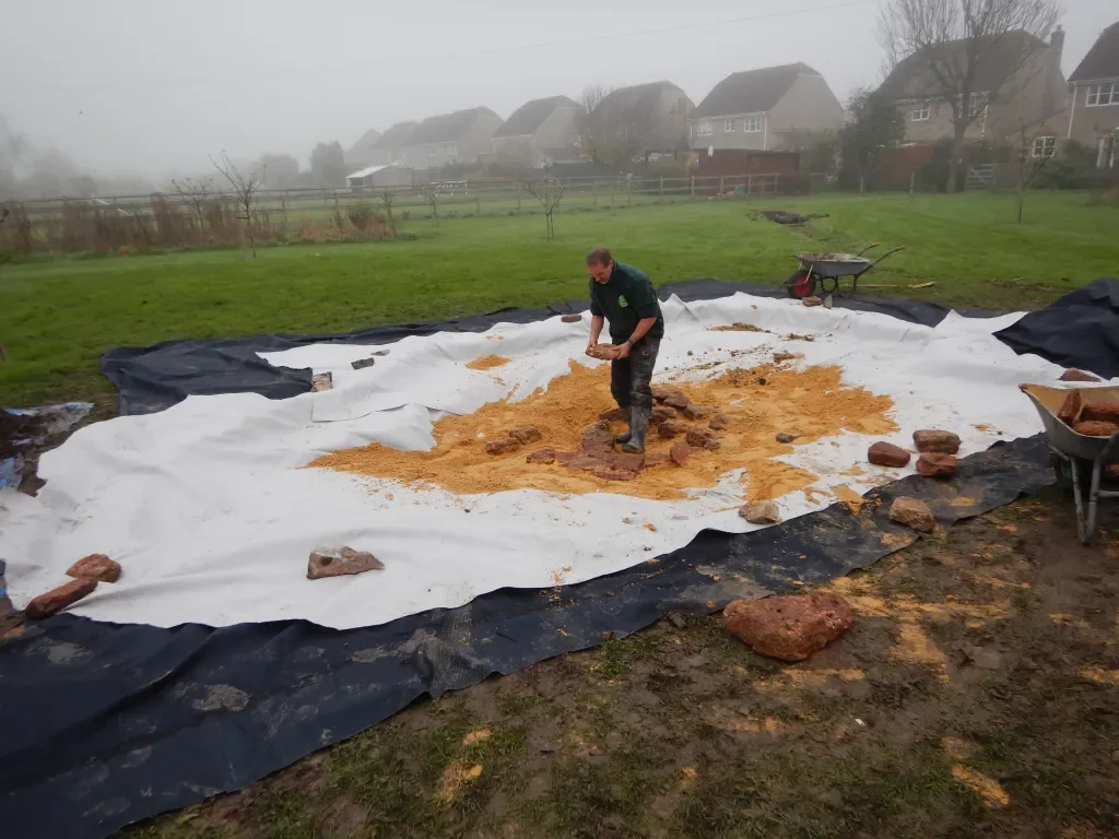 A person working on a large outdoor project, spreading or leveling material on a fabric ground covering, with a wheelbarrow and other tools nearby in a yard on a cloudy day.