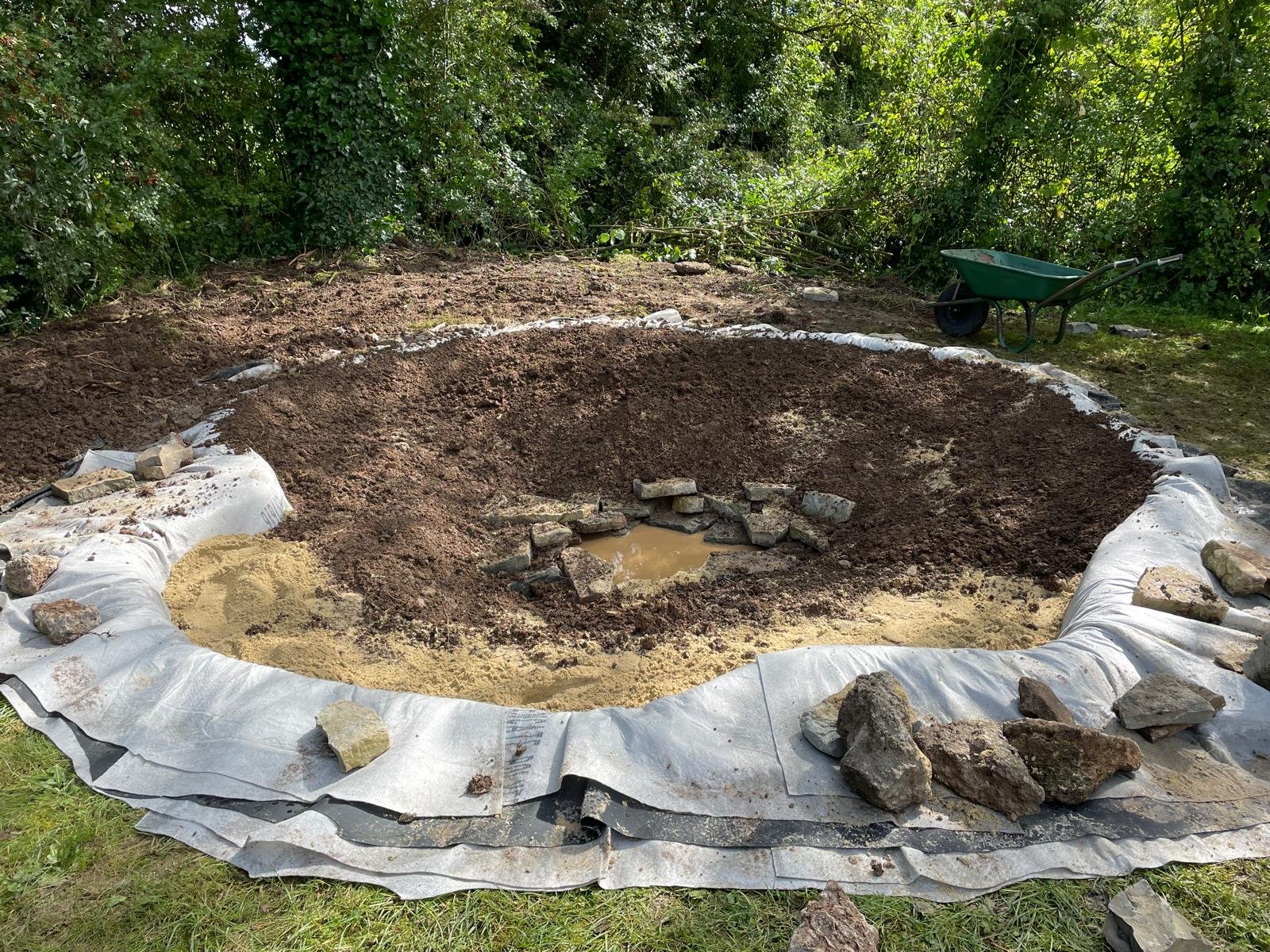 A large hole in the ground surrounded by a white geotextile fabric, with rocks on the edge, set in a grassy outdoor area with green bushes and trees in the background, and a green wheelbarrow nearby.