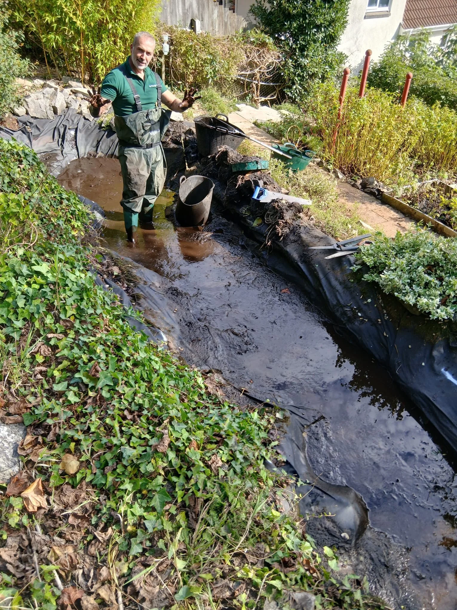 Tony Watt, The Nature Gardener working on a wildlife pond project, standing in a muddy area with a garden pond, surrounded by plants and greenery, holding muddy gloves and wearing garden work clothes.