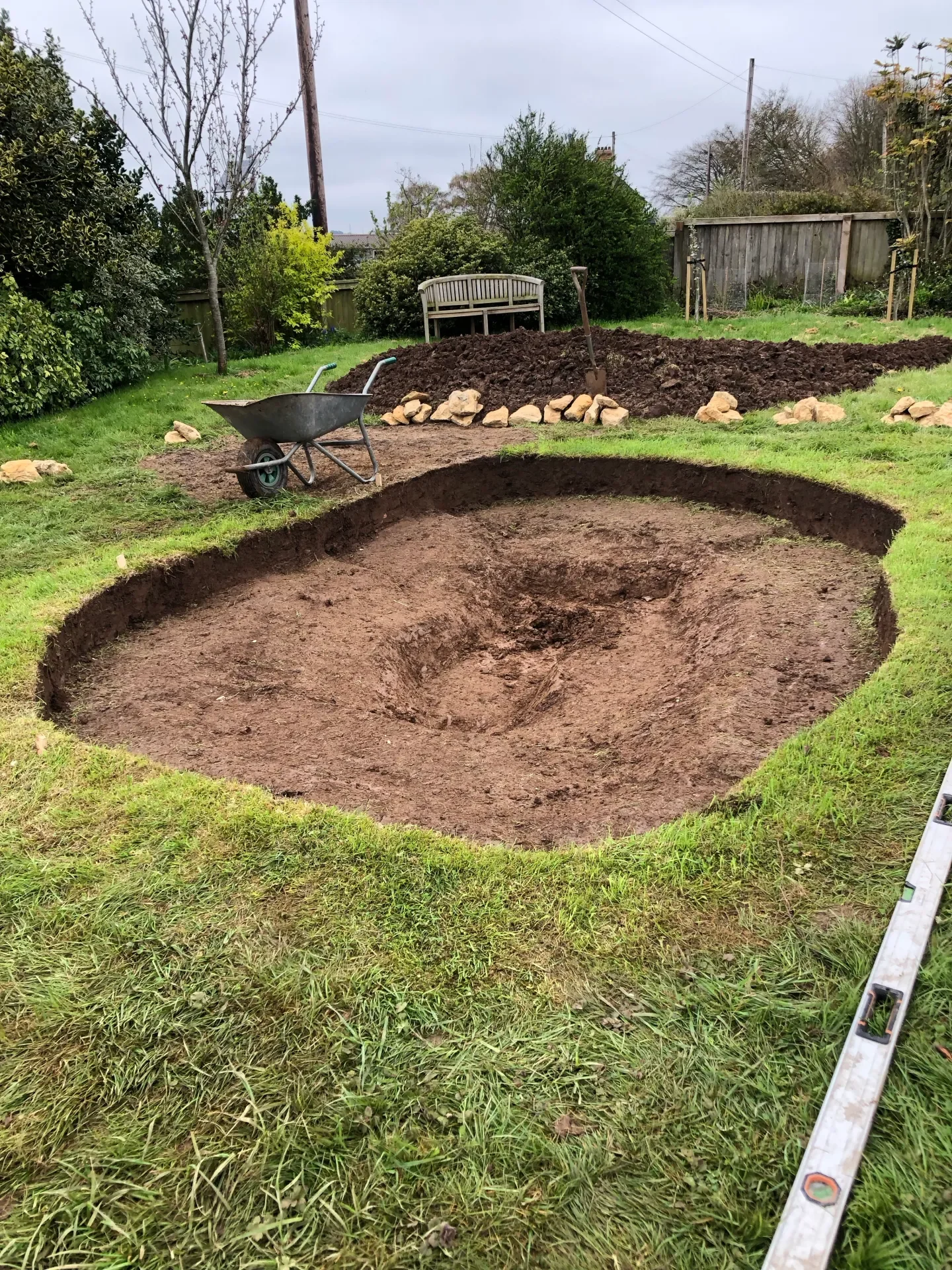 A garden with a large dug hole, a wheelbarrow, and a shovel, surrounded by grass and trees, with a wooden fence in the background.