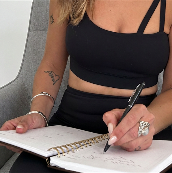Close-up of a woman wearing a black crop top with double straps, writing in a spiral notebook with a black pen.