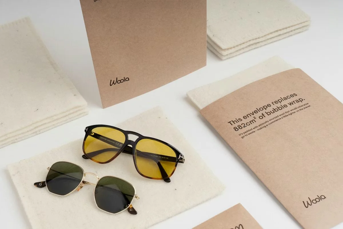 Two pairs of sunglasses, one with yellow-tinted lenses and black frame, the other with dark lenses and a gold frame, along with beige and brown packaging materials, including a cardboard box and envelopes.