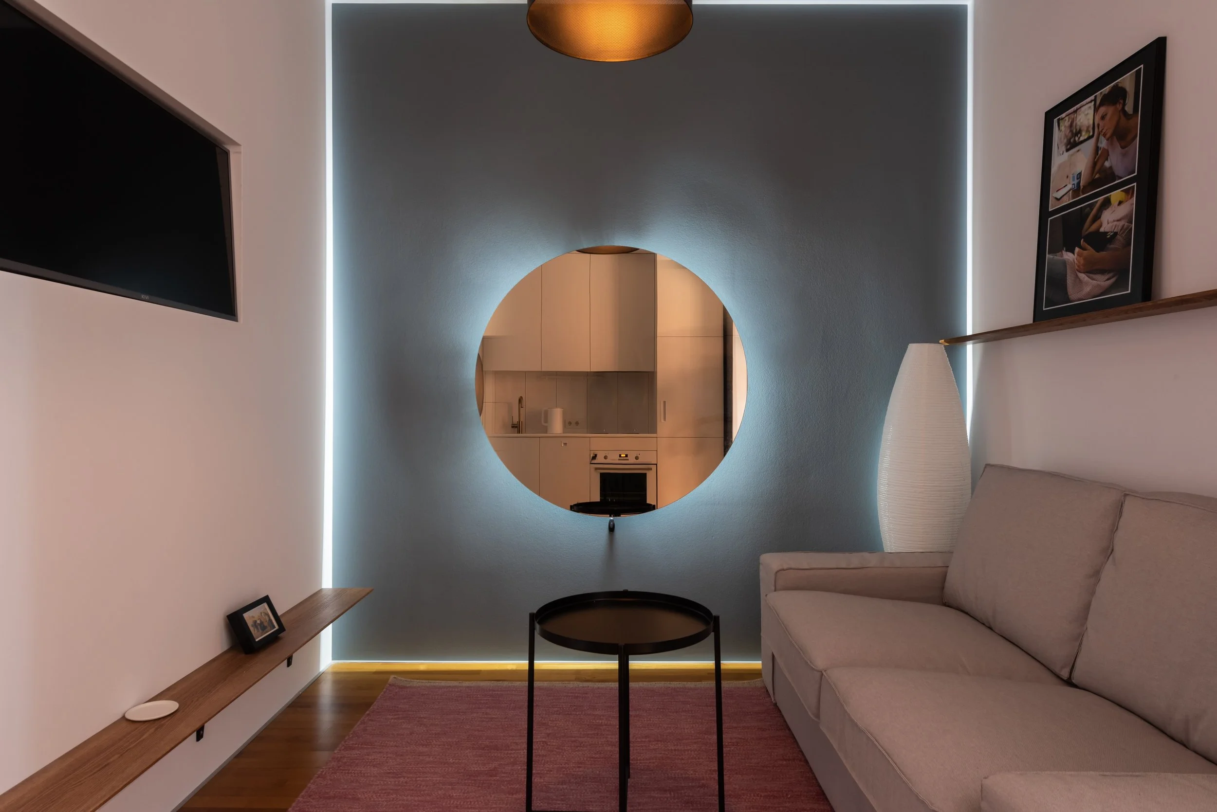 Un salon moderne avec un mur bleu clair, un canapé beige, une table ronde noire, une lampe blanche, un tableau photo sur le mur et une télévision.
