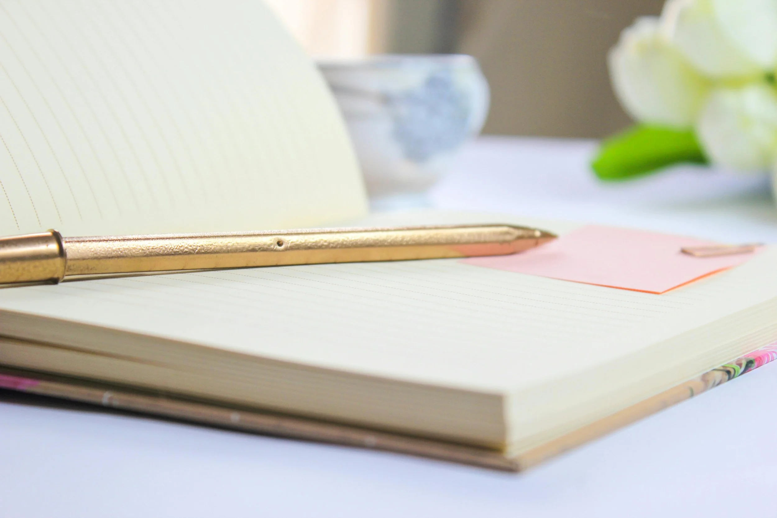Une pile de cahiers ou de blocs-notes ouverts avec un stylo appuyé dessus, un bol en céramique et des fleurs blanches floues en arrière-plan.