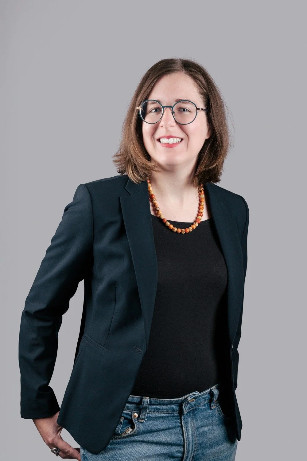 Femme souriante portant des lunettes, un blazer noir, un collier en perles, et un jean, posant contre un fond gris.