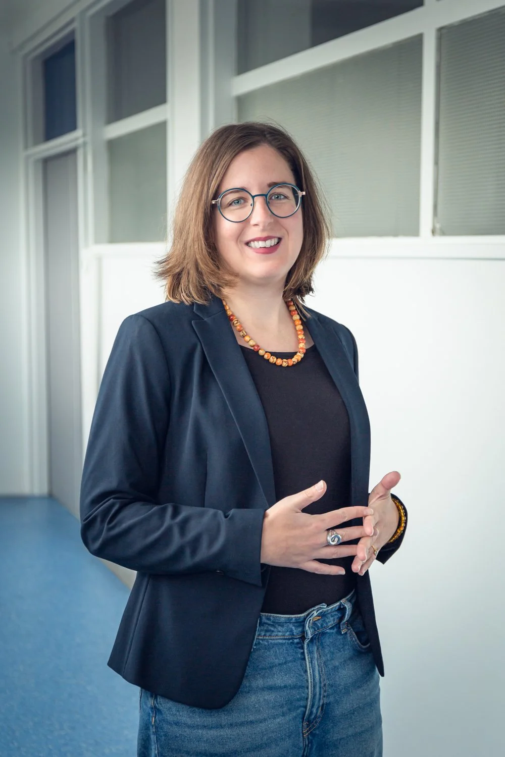 Une femme souriante portant des lunettes, un blazer noir, un t-shirt noir, un collier orange, un bracelet et une bague, debout dans un environnement intérieur moderne avec des fenêtres.
