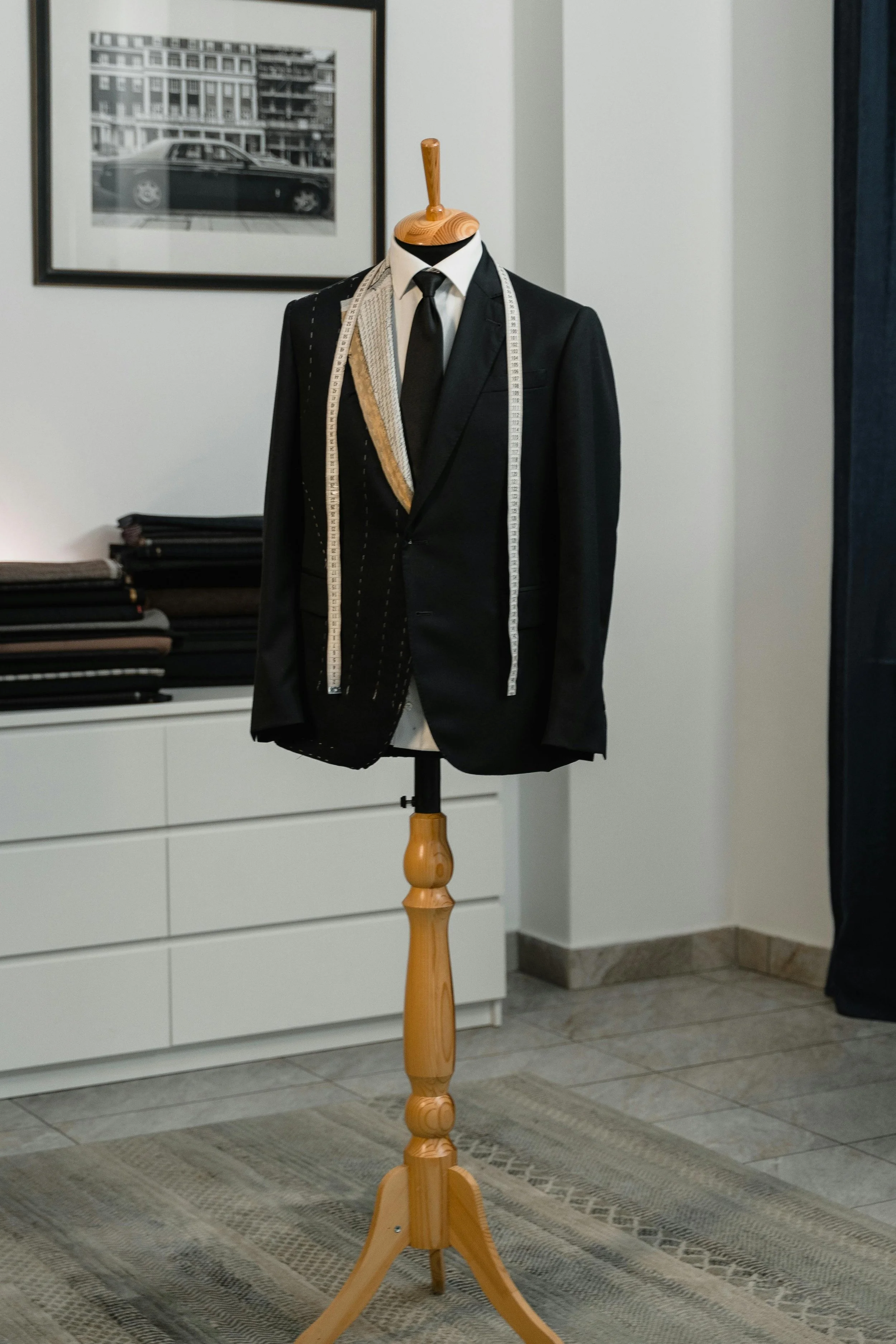Mannequin habillé d'un costume noir avec une chemise blanche, une cravate noire, portant un mètre ruban autour du cou, sur un support en bois, dans une pièce avec des armoires blanches et un tableau noir et blanc au mur.